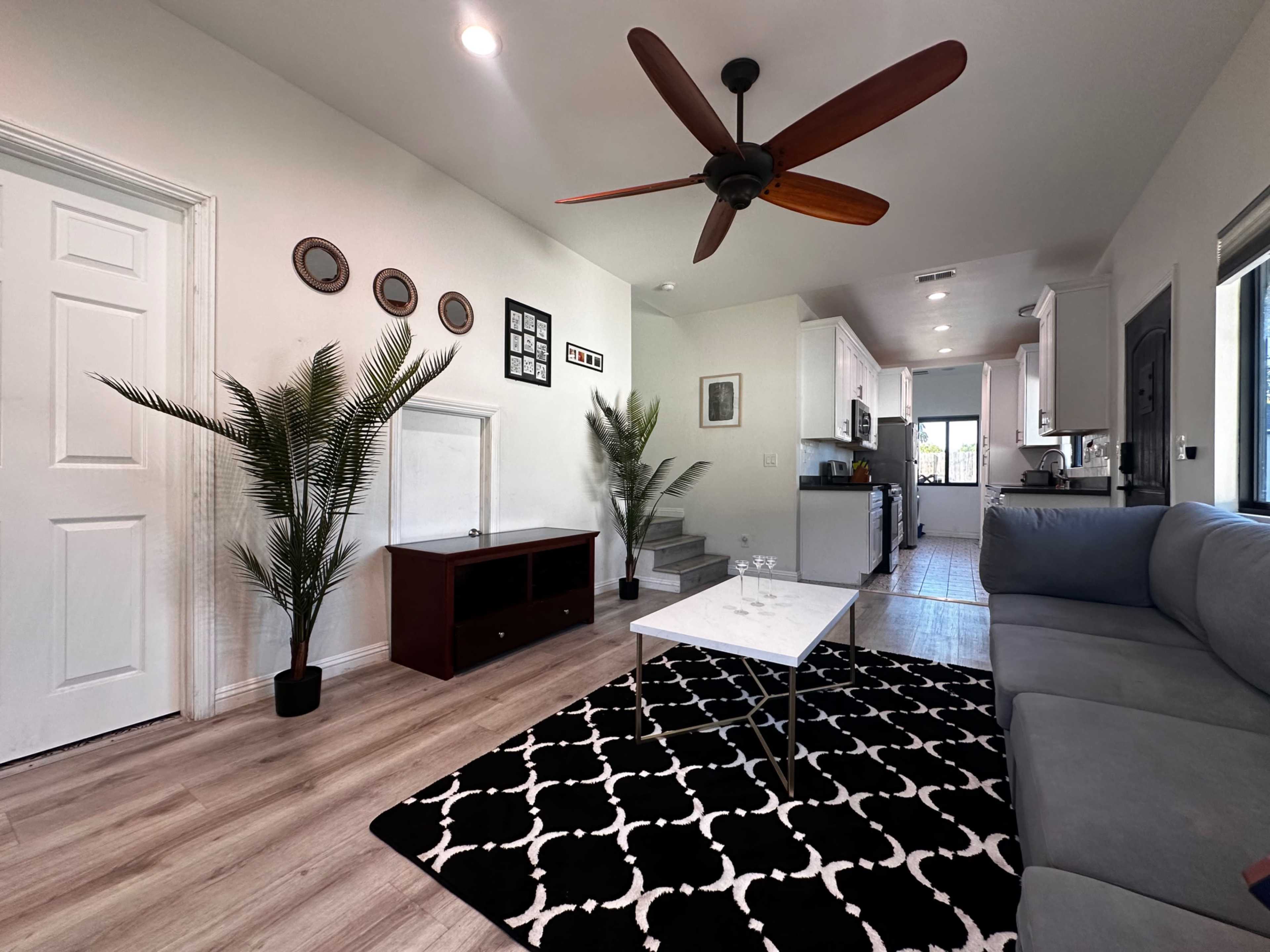 A modern living room with a ceiling fan, a black and white patterned rug, a coffee table, potted plants, and a view of a kitchen area in the background.