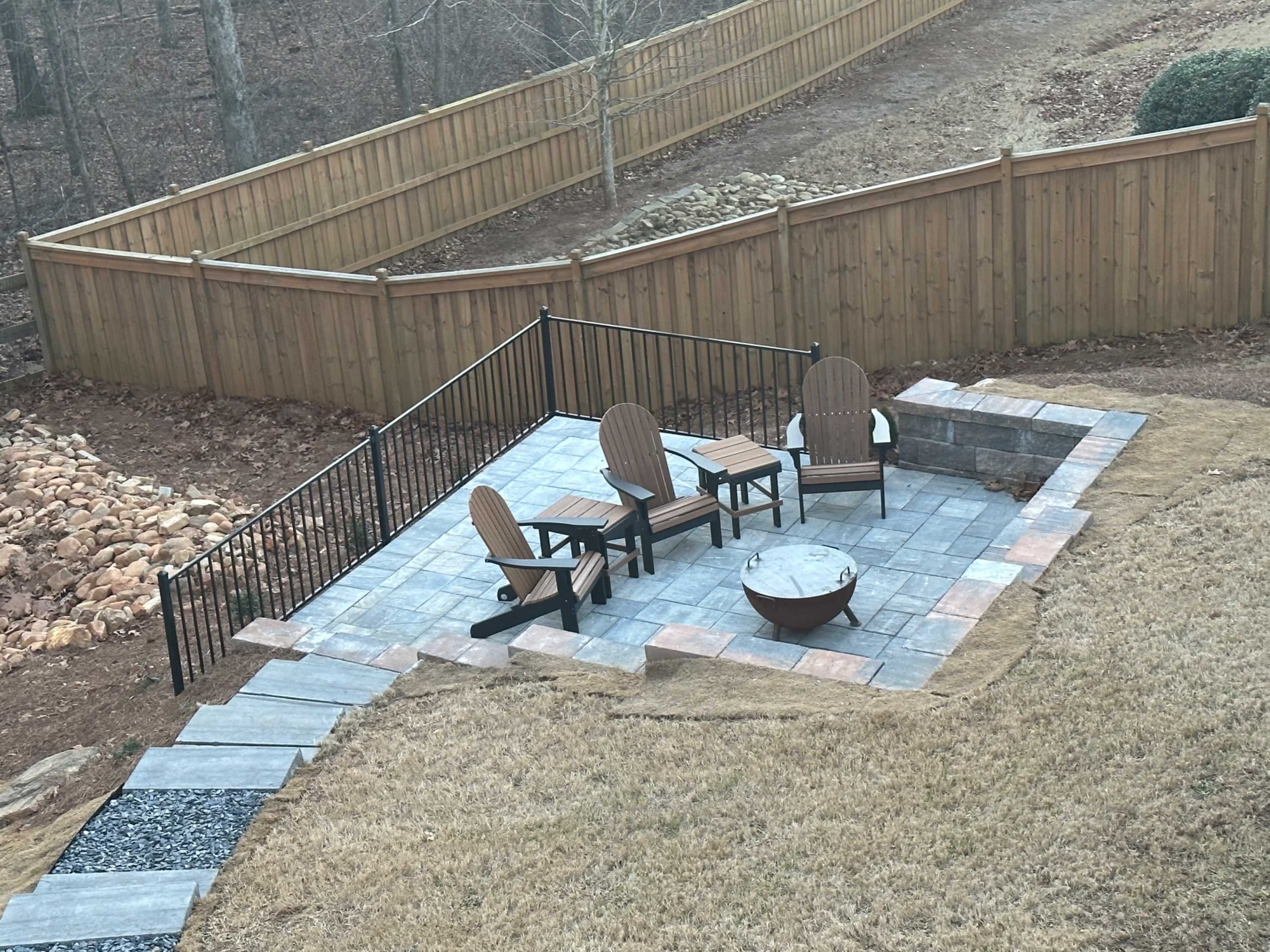 A patio area with three wooden chairs arranged around a circular fire pit, bordered by a stone walkway and surrounded by a wooden fence.