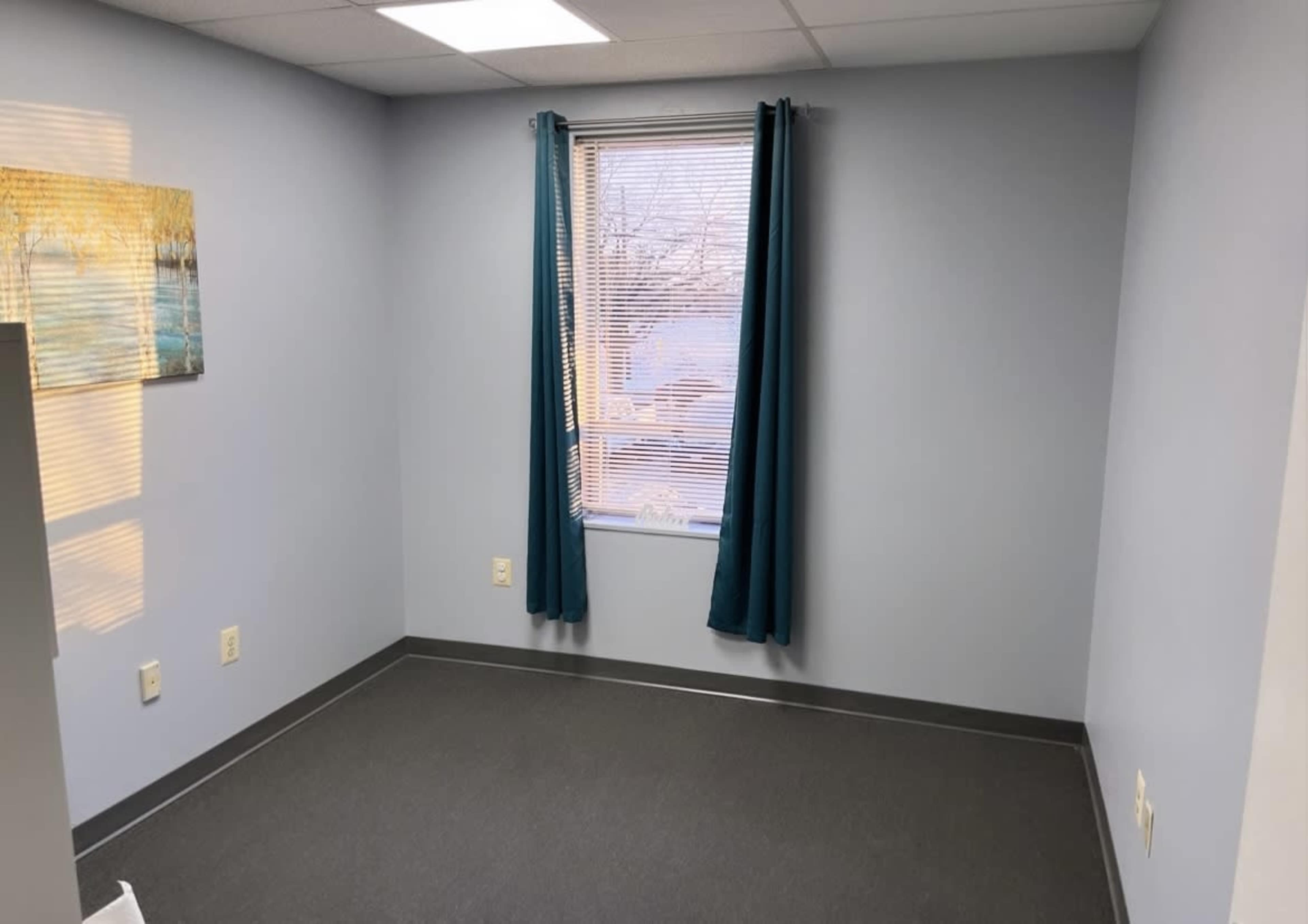 A sparsely furnished room with light blue walls, a window covered by teal curtains, and a single piece of artwork on the wall.