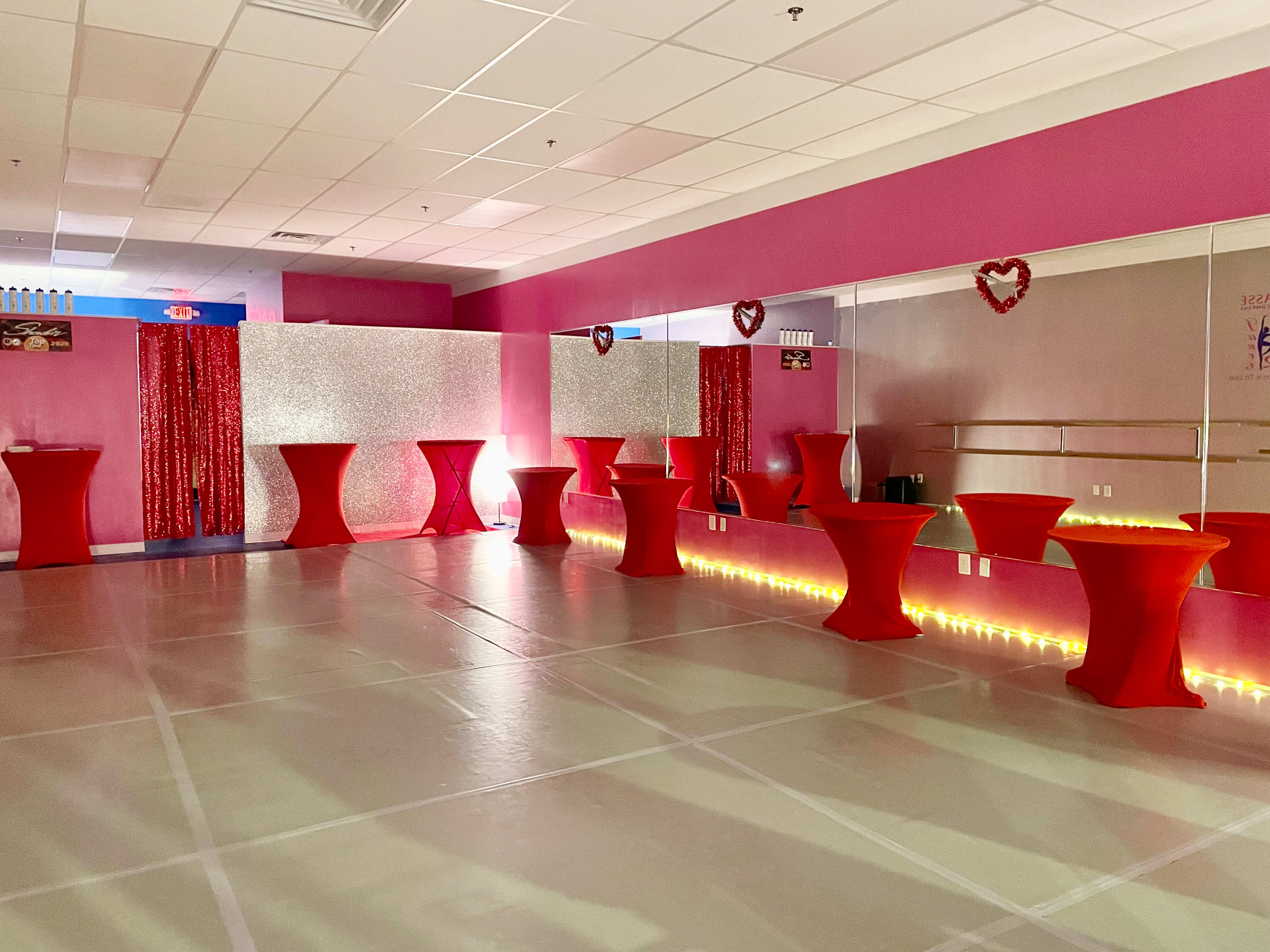 The image shows an event space featuring red tablecloth-covered tables, pink walls, and decorative lighting along the floor.