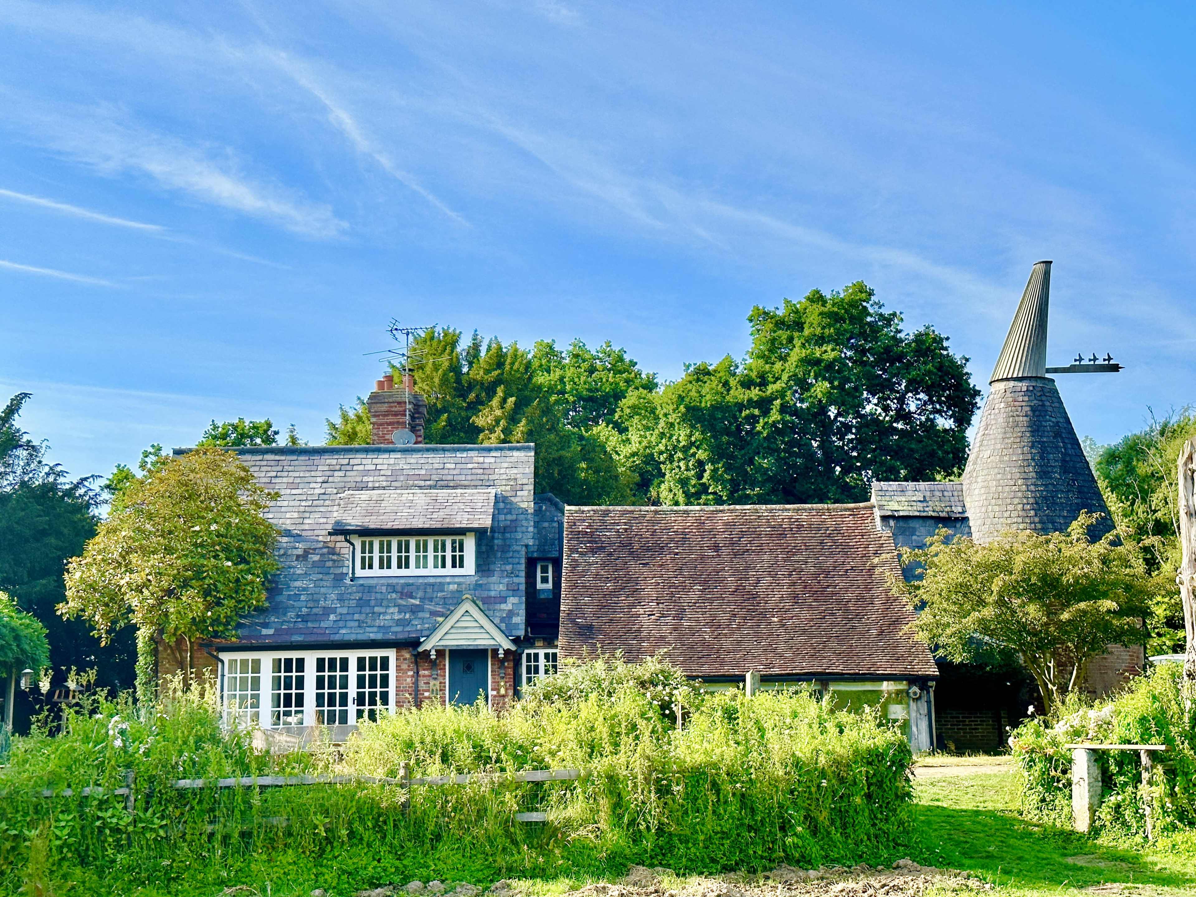A quaint house with a conical roof structure surrounded by lush greenery and trees under a clear blue sky.