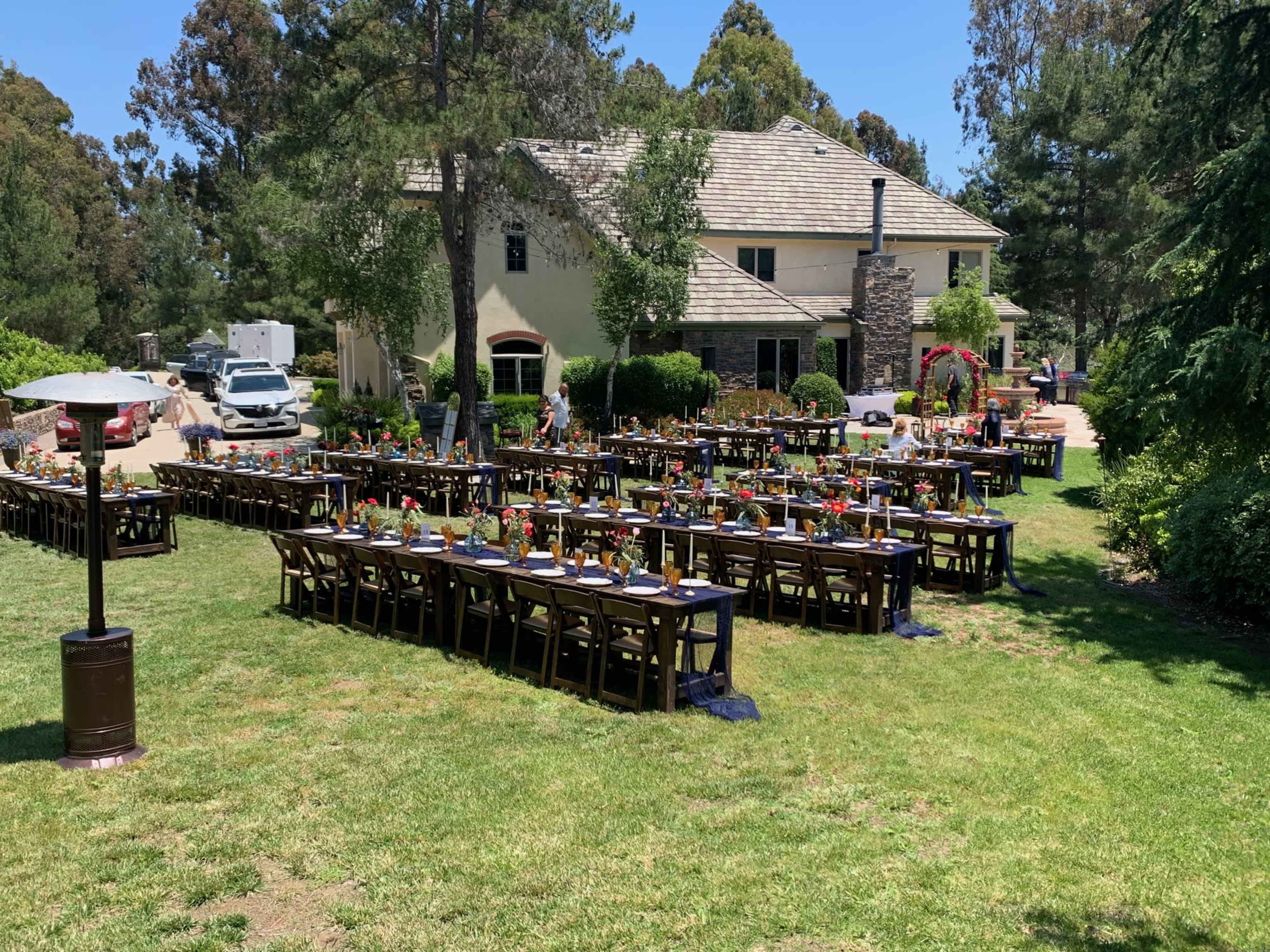 A large outdoor dining setup with several long tables is arranged on a grassy area in front of a spacious house, surrounded by trees.