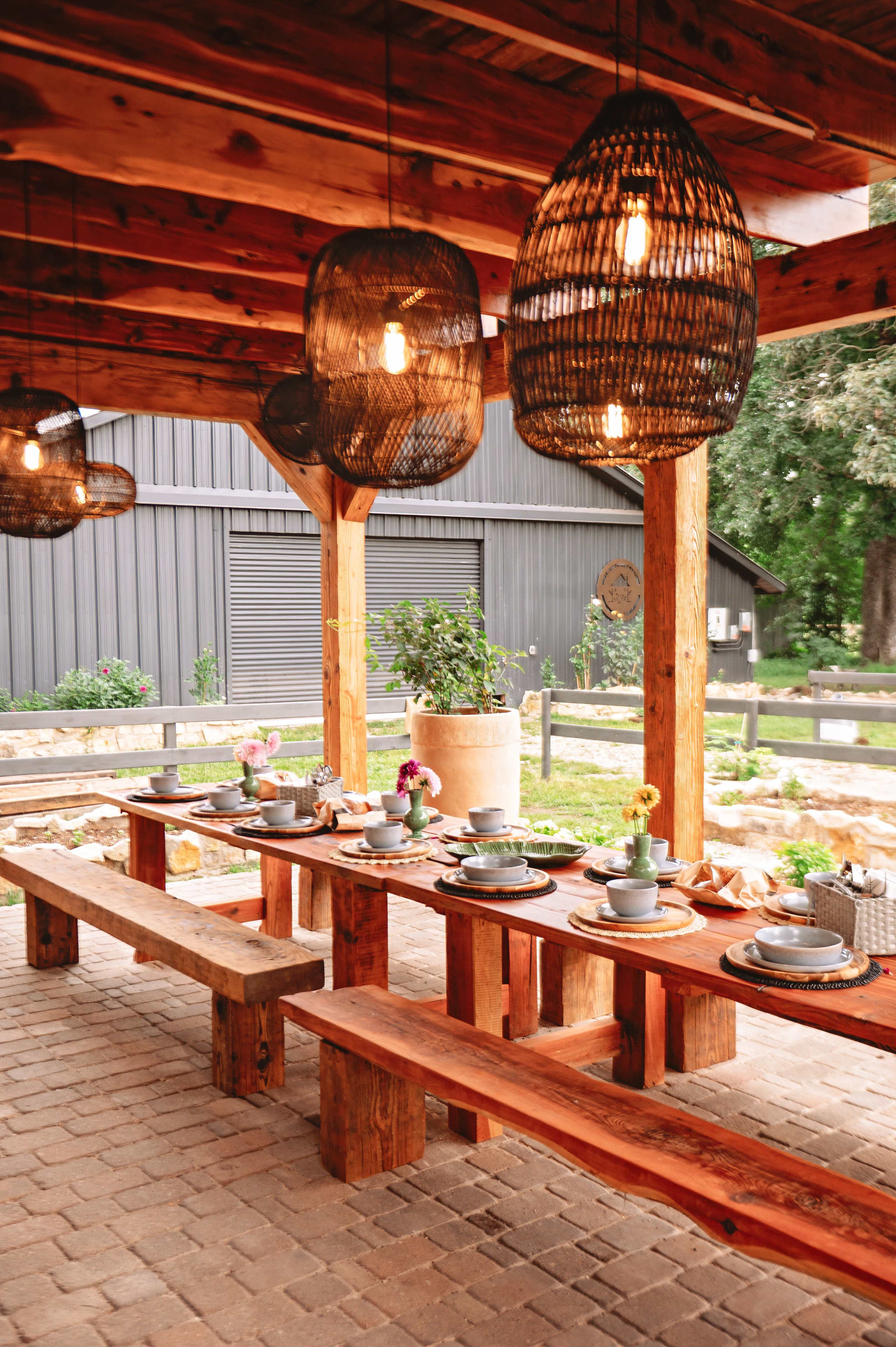 A rustic outdoor dining area features a long wooden table set with dishes and surrounded by woven pendant lights.