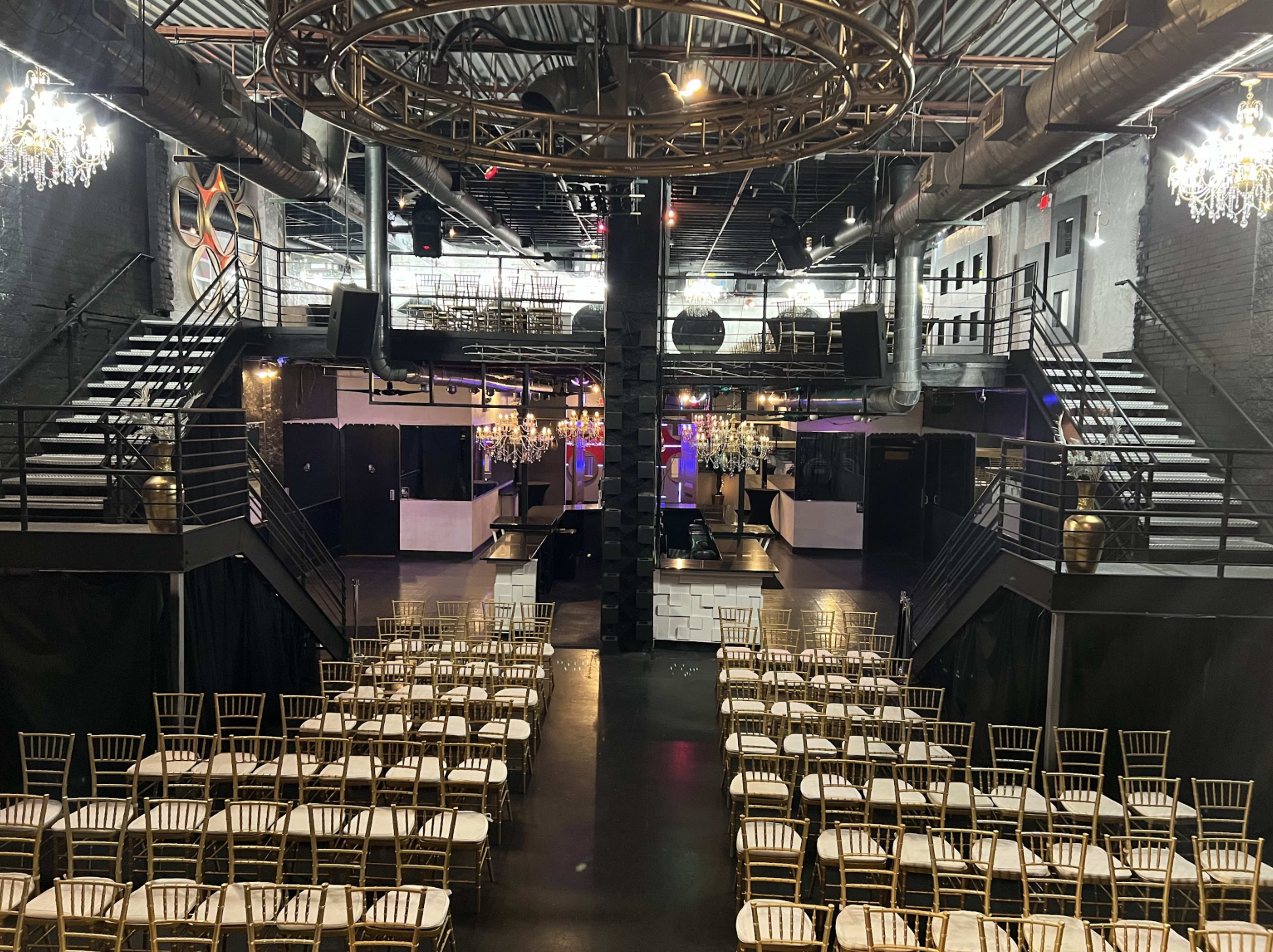 A spacious event venue with rows of gold chairs arranged in front of a stage, flanked by two staircases and adorned with chandeliers.
