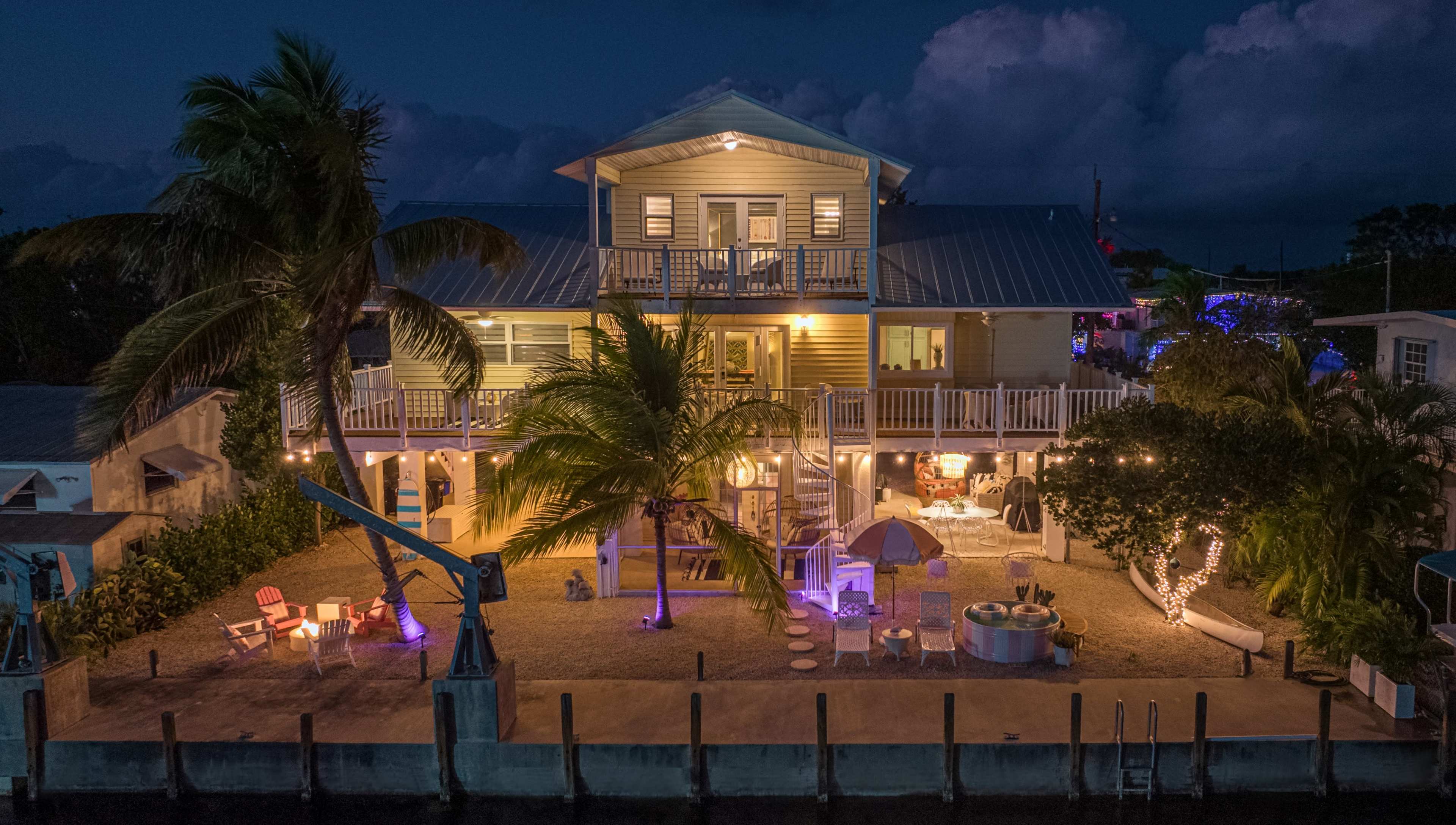 A two-story waterfront house with a balcony and outdoor seating is illuminated by lights at night, surrounded by palm trees.