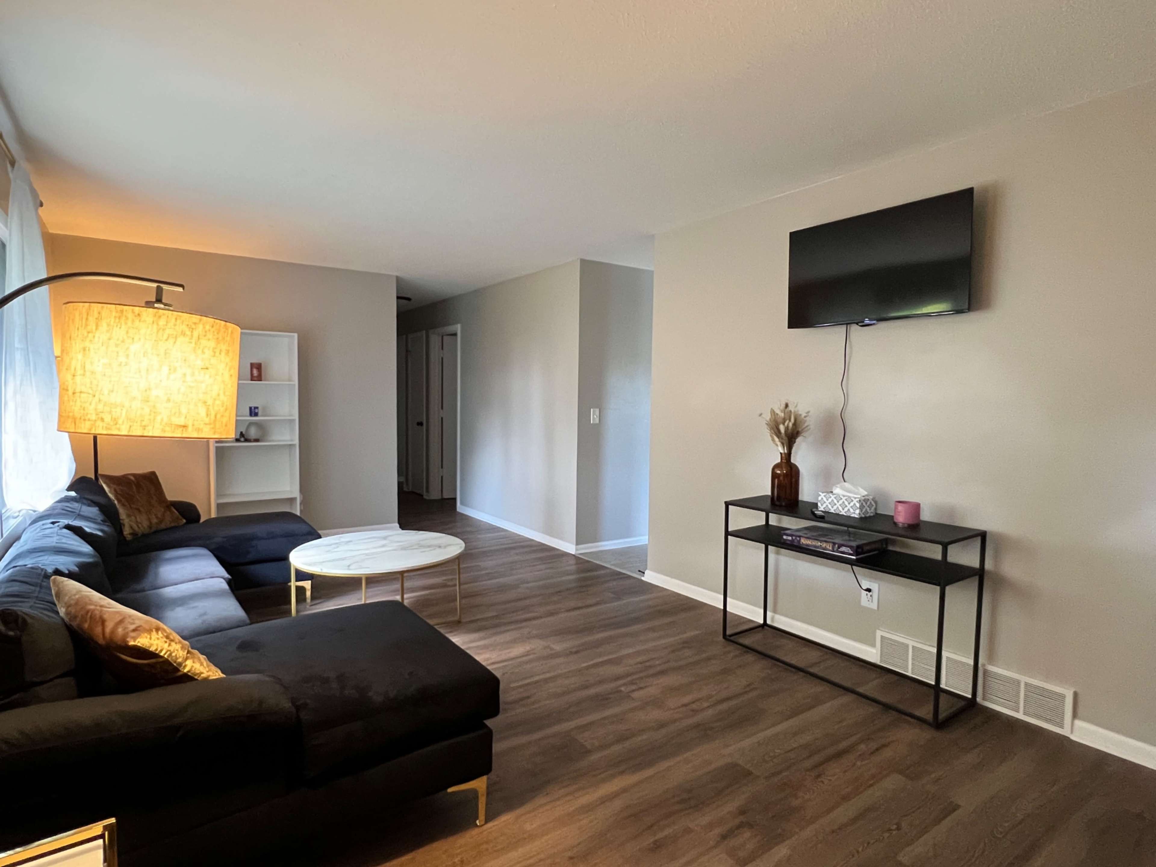 A living room with a sectional couch, a round coffee table, a television mounted on the wall, and a light fixture.
