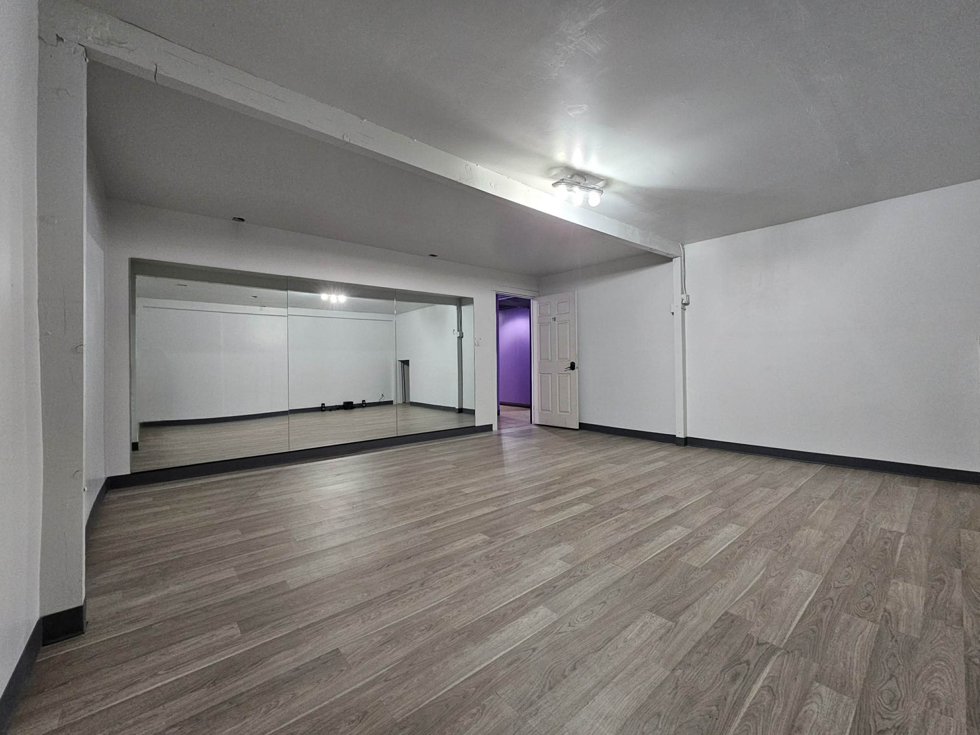 The image shows a spacious, empty room with light-colored wooden flooring and a mirrored wall, featuring a door that leads to a purple wall.