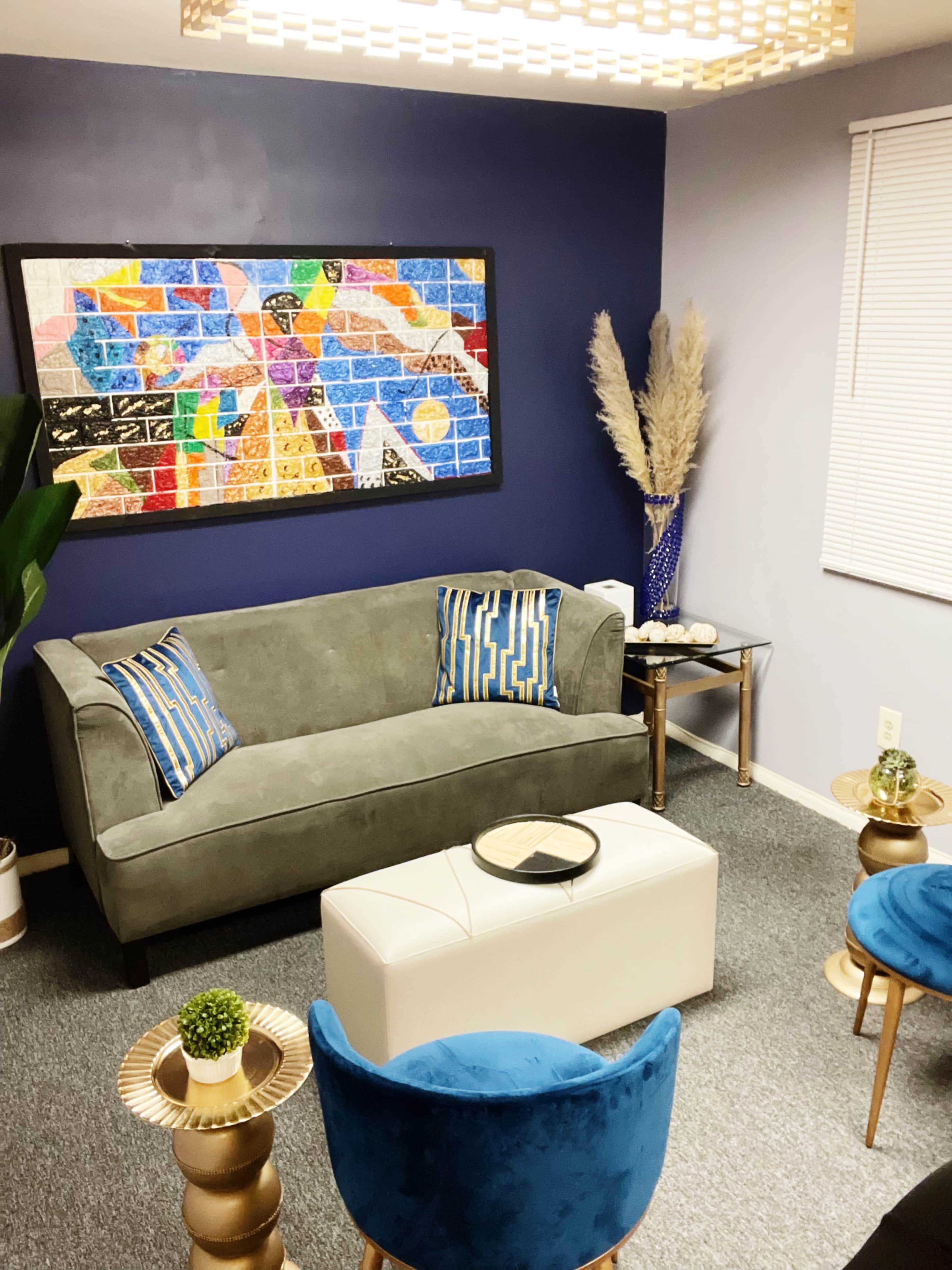 A modern living room features a gray sofa, two blue chairs, a white ottoman, and a colorful wall art piece against a dark blue wall.