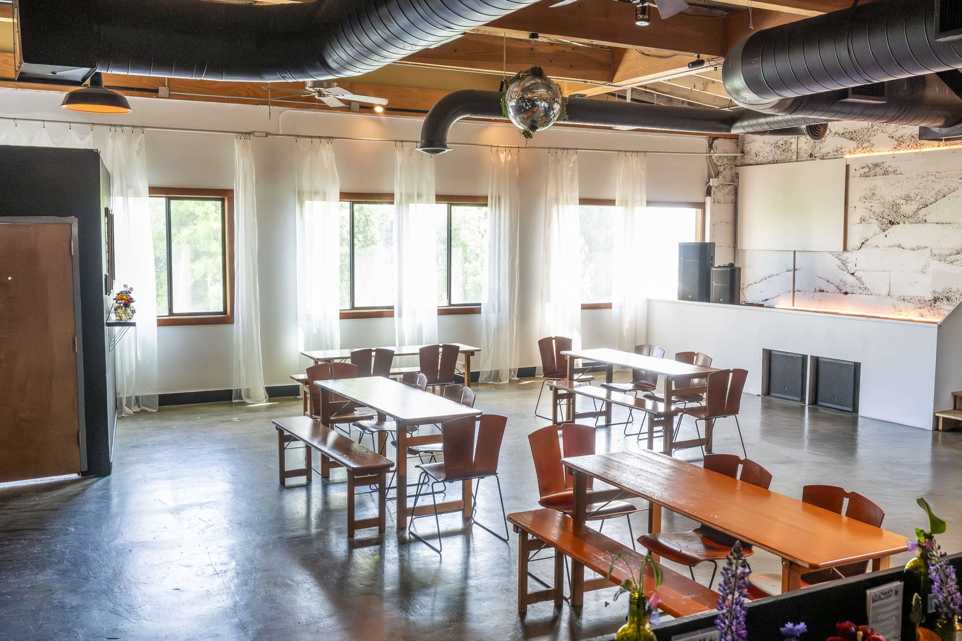 The image shows a spacious interior of a dining area featuring several wooden tables and benches, large windows with sheer curtains, and a minimalist bar counter at one end.