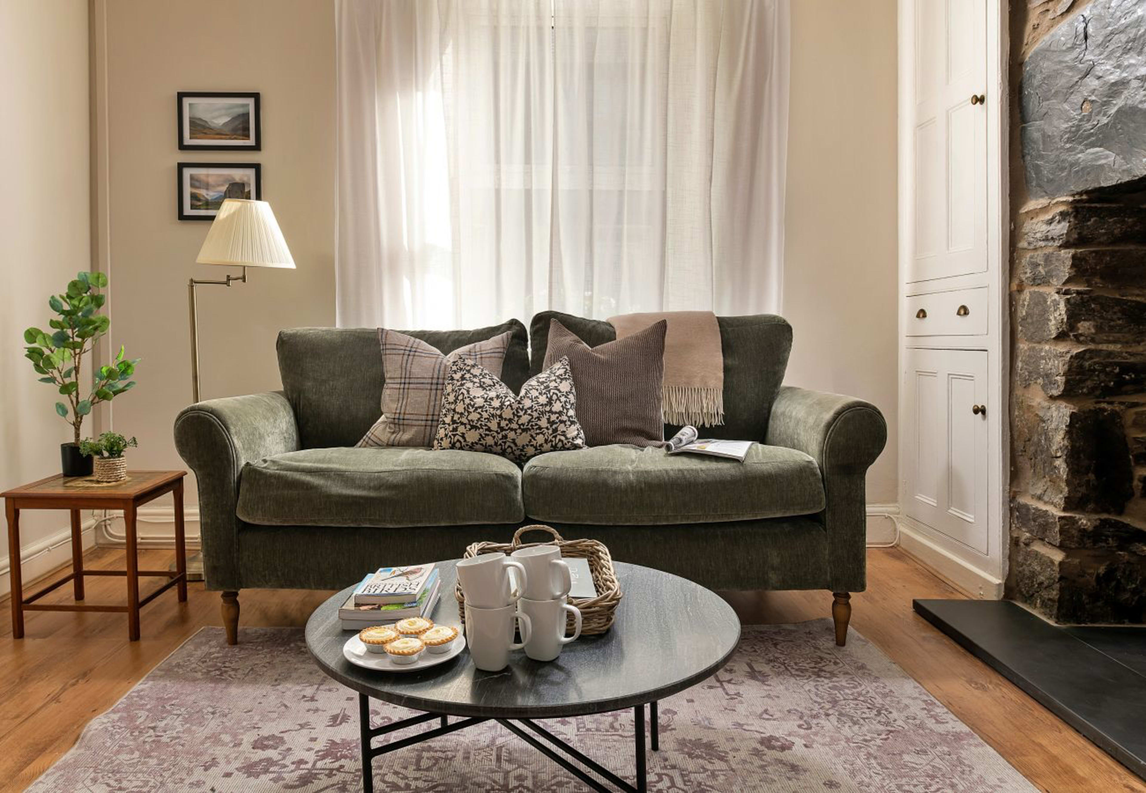 A green sofa with patterned pillows is paired with a coffee table holding a tea set and pastries, set in a cozy room with light curtains and a stone fireplace.