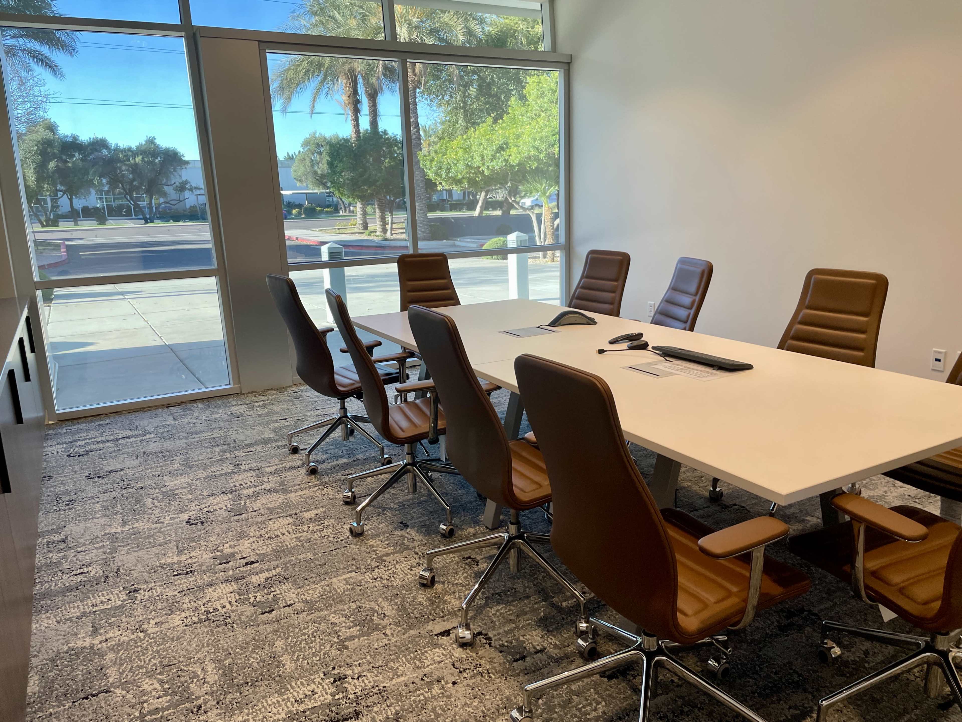A conference room features a large white table surrounded by brown leather chairs, with large windows allowing natural light to enter.
