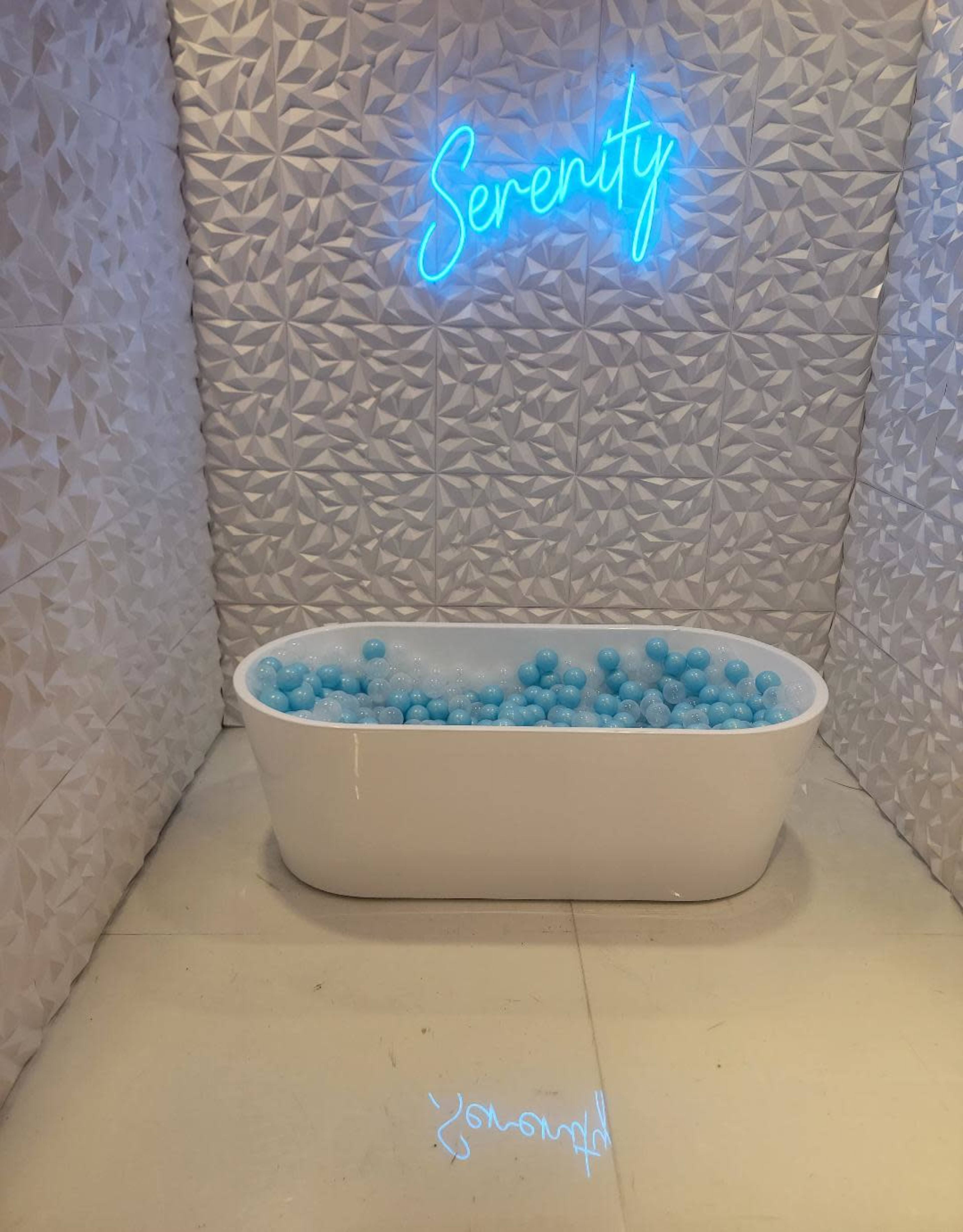 A white bathtub filled with blue balls sits beneath a neon sign reading "Serenity" against a textured white wall.