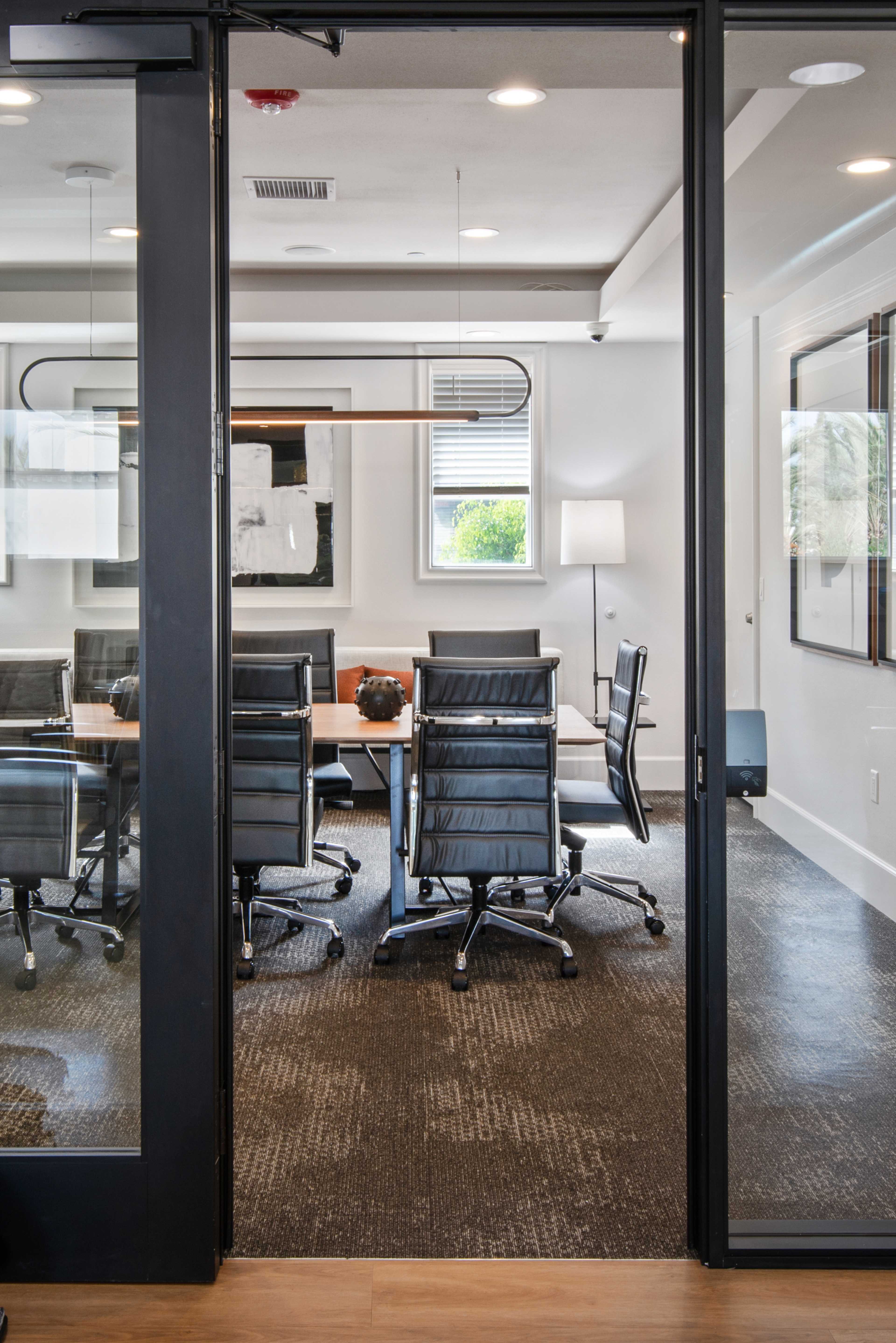 A modern conference room featuring a long wooden table surrounded by black rolling chairs, with large windows allowing natural light to enter.