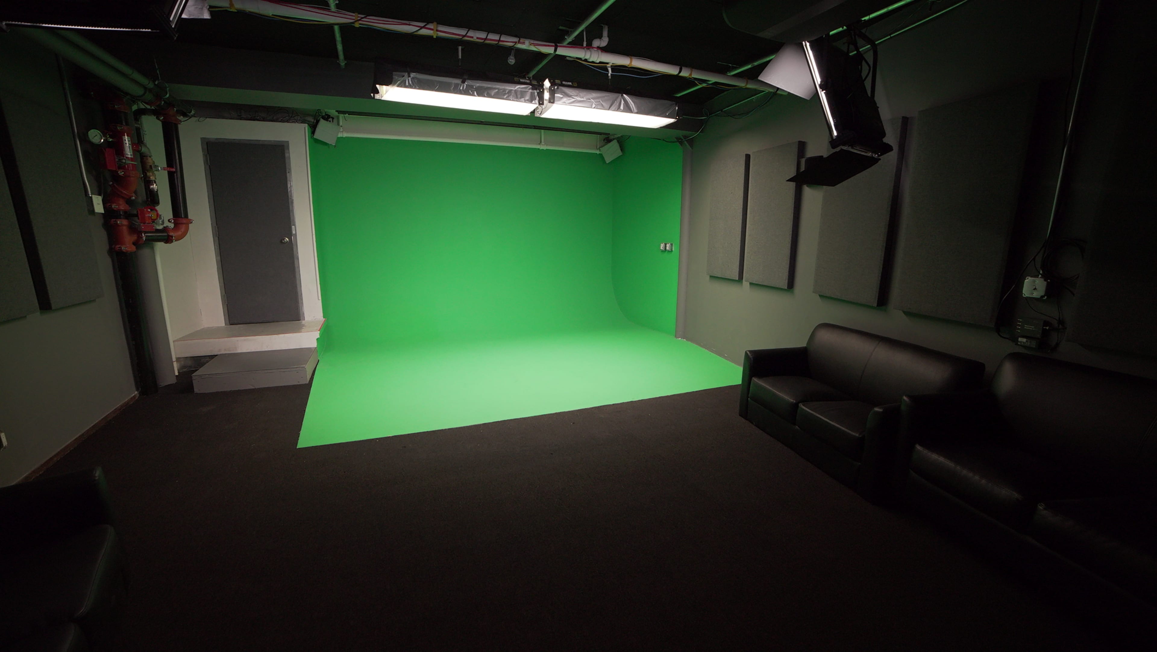 A green screen studio with a green backdrop, a door, soundproof panels, and two black couches.