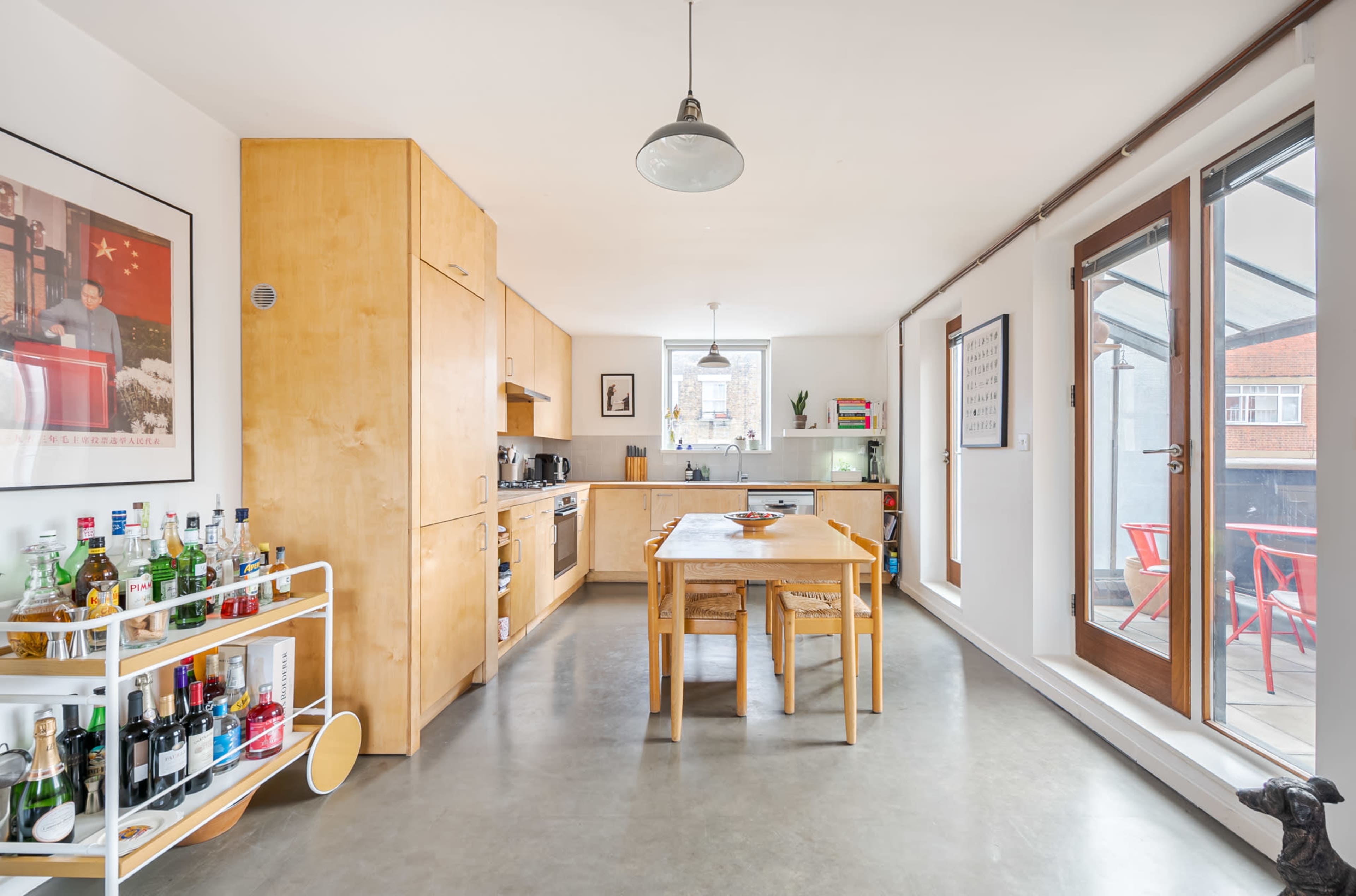A modern kitchen features wooden cabinetry, a central dining table, and a bar cart filled with various beverages, with large windows providing natural light.
