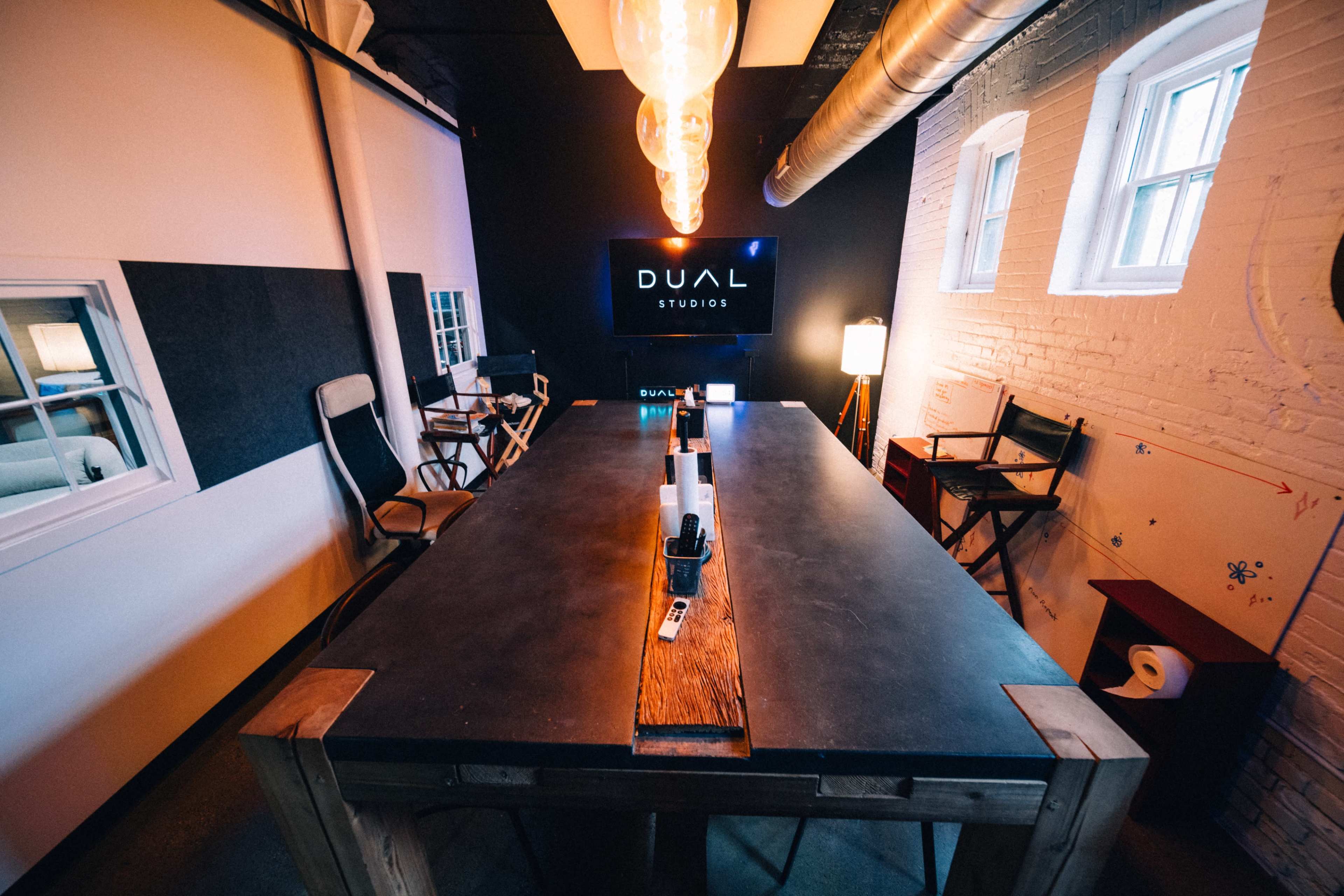 The image shows a modern conference room with a long wooden table, director's chairs, and a wall-mounted screen displaying the name "DUAL STUDIOS."