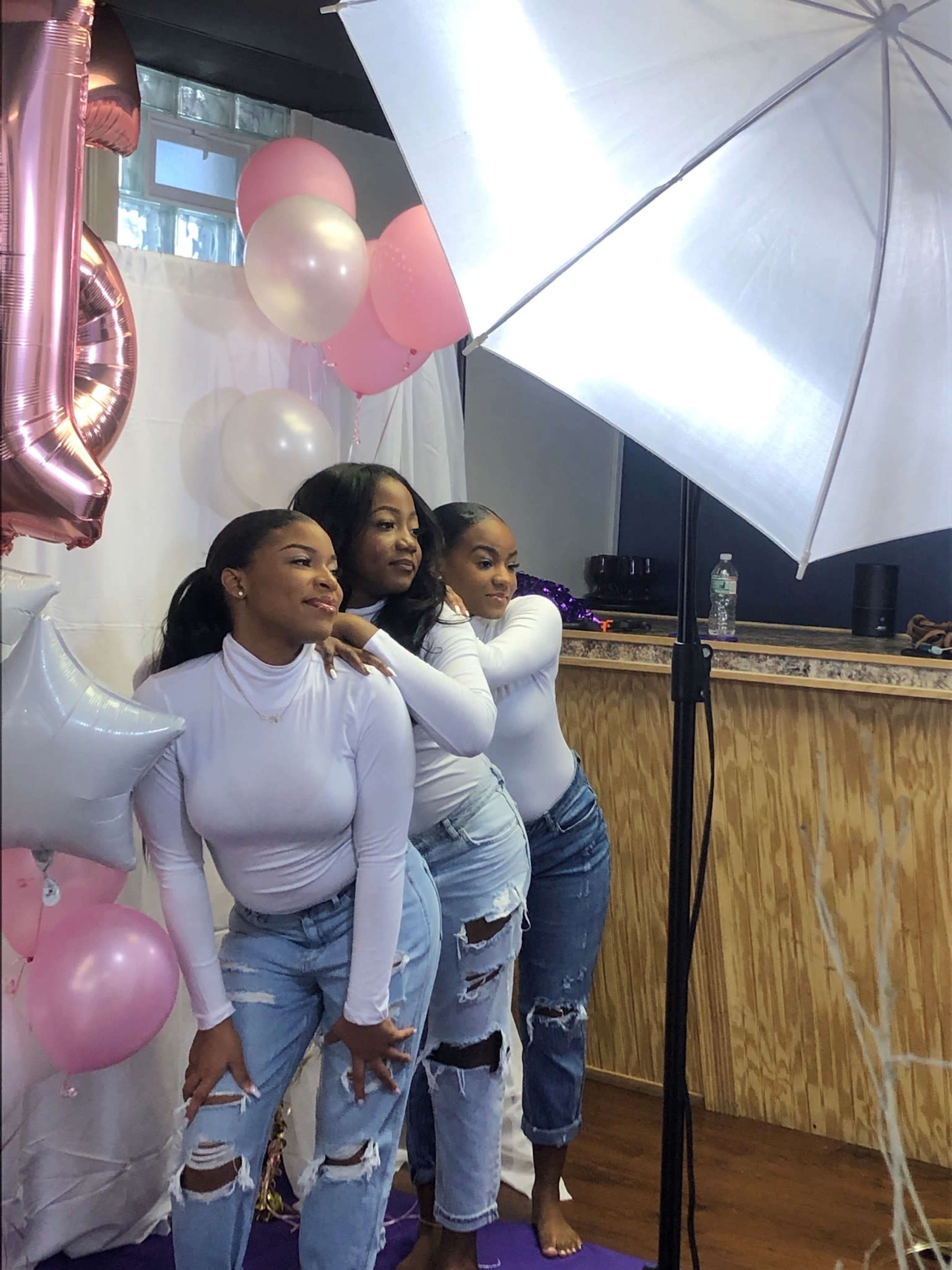 Three women pose together against a backdrop of balloons and decorations, dressed in white tops and distressed jeans, with a lighting umbrella nearby.