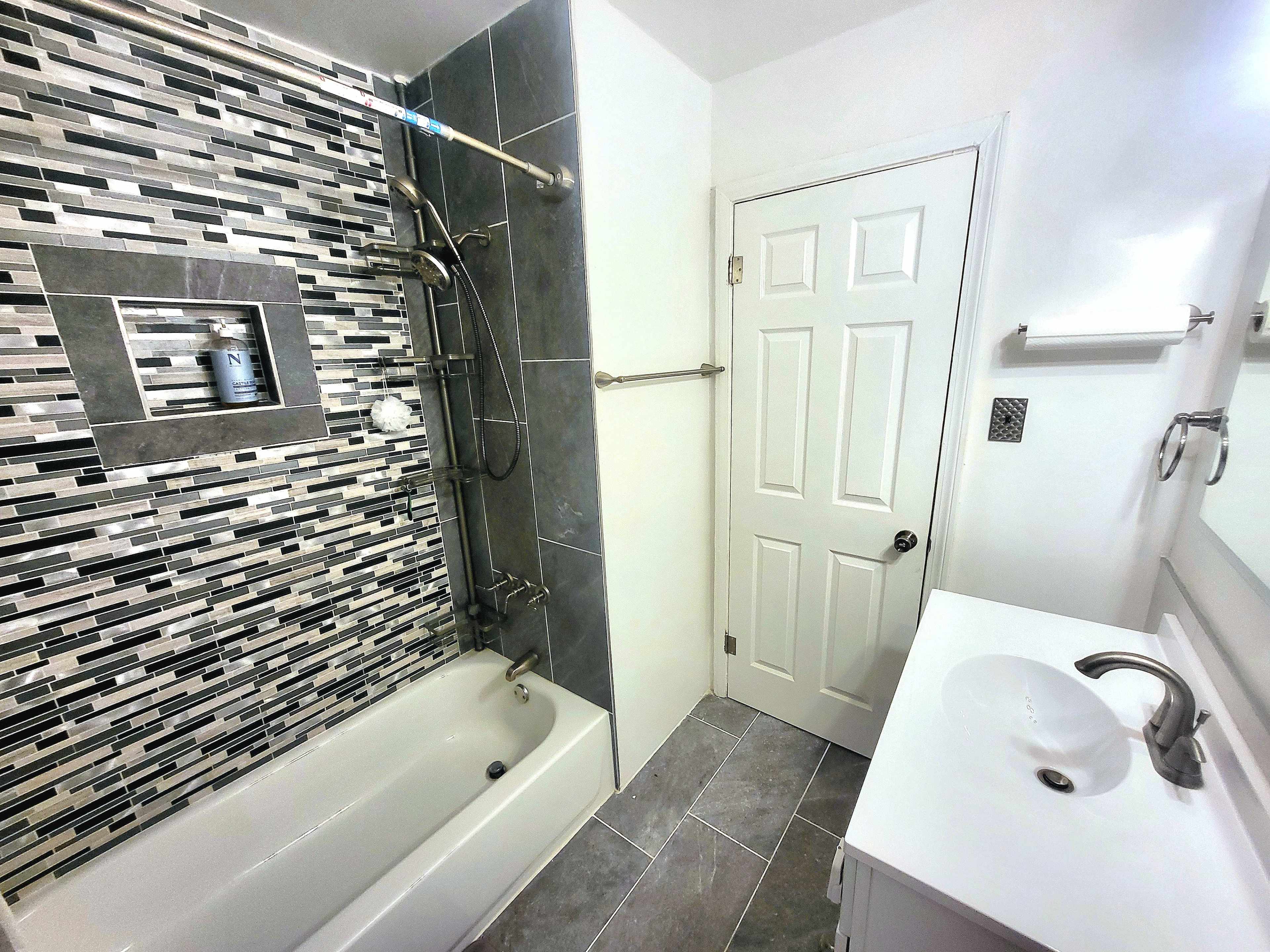 The image shows a modern bathroom featuring a bathtub with a tiled surround, a sleek sink, and a door leading to another area.
