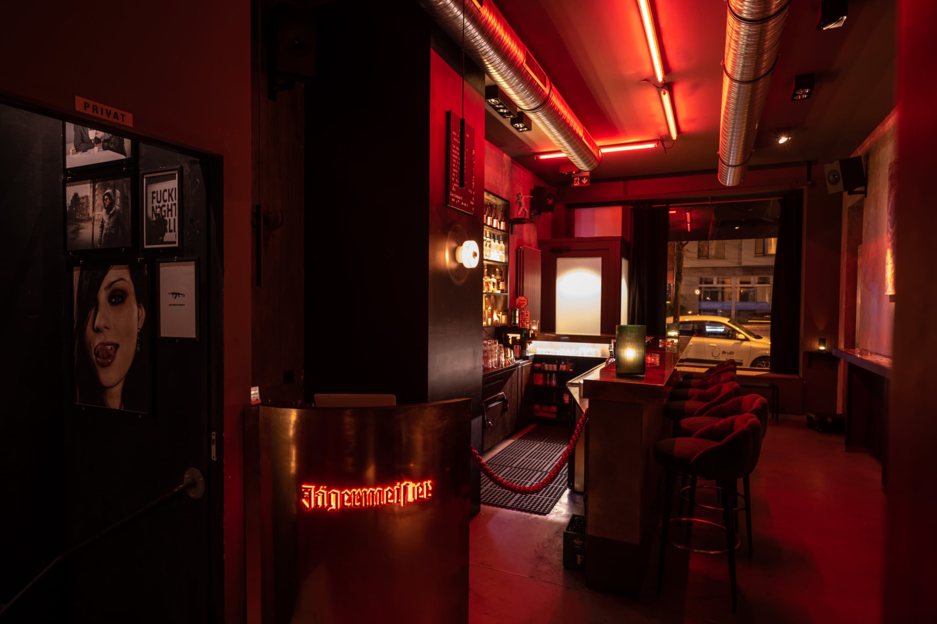 The image shows a dimly lit bar with red neon lighting, featuring a counter with stools and a selection of bottles displayed on shelves.