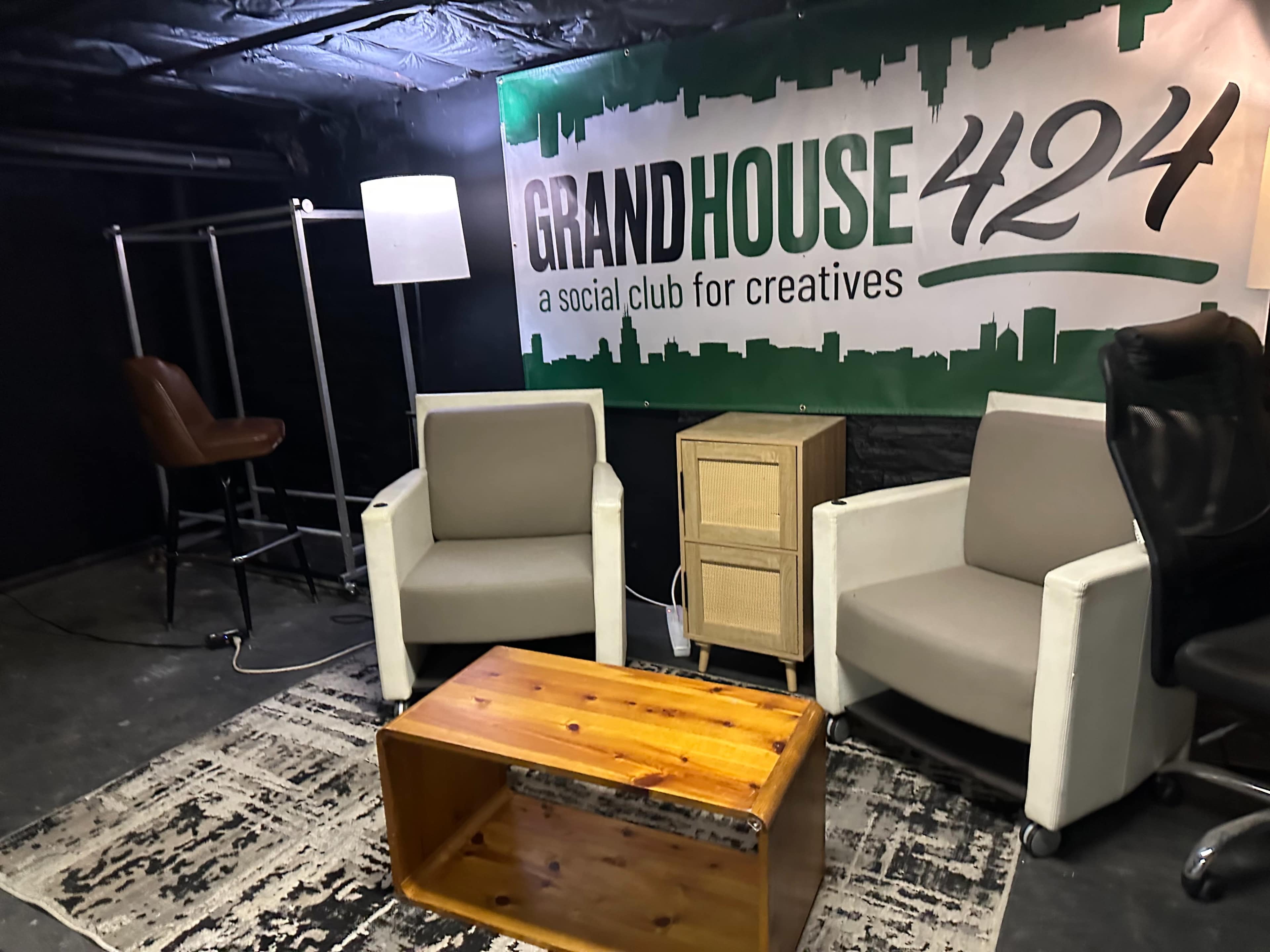 The image shows a small, dimly lit lounge area with two white armchairs, a wooden coffee table, a floor lamp, a tall clothing rack, and a sign that reads "GRANDHOUSE 424."