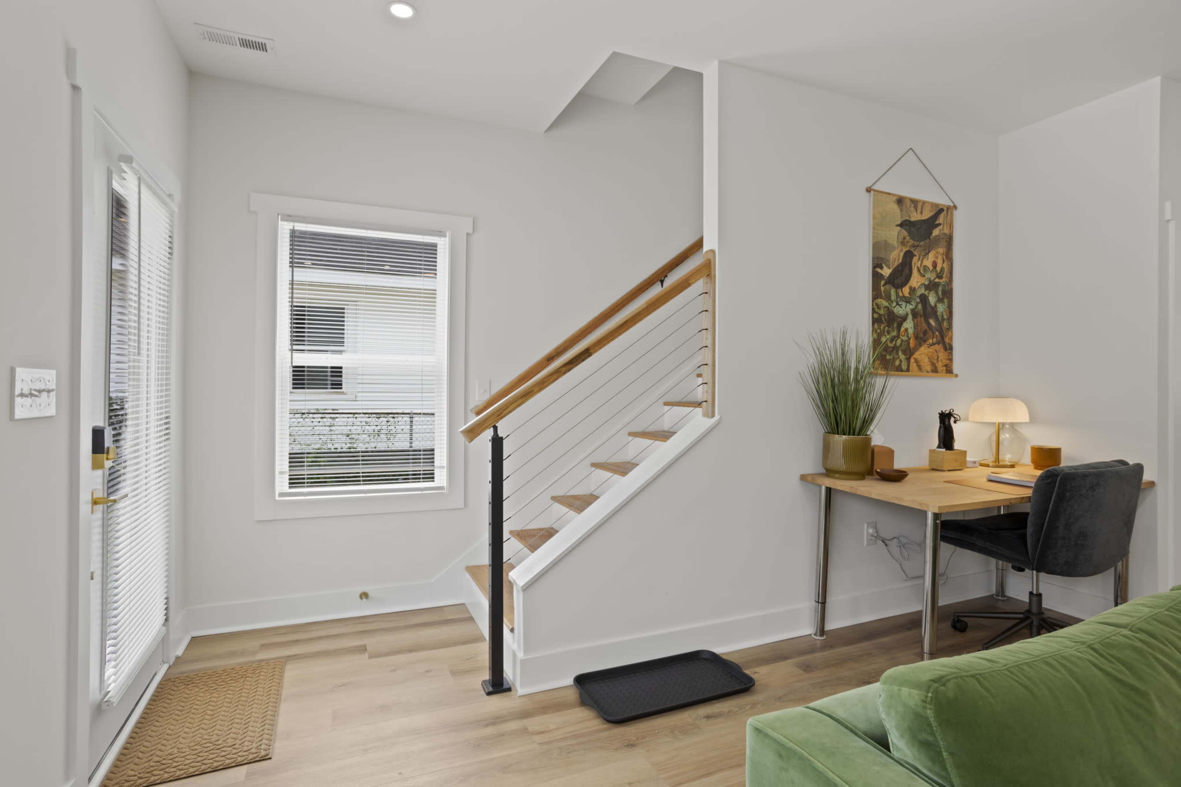 The image shows a modern entryway with a staircase, a small desk with a lamp, and a green couch in a bright, airy space.