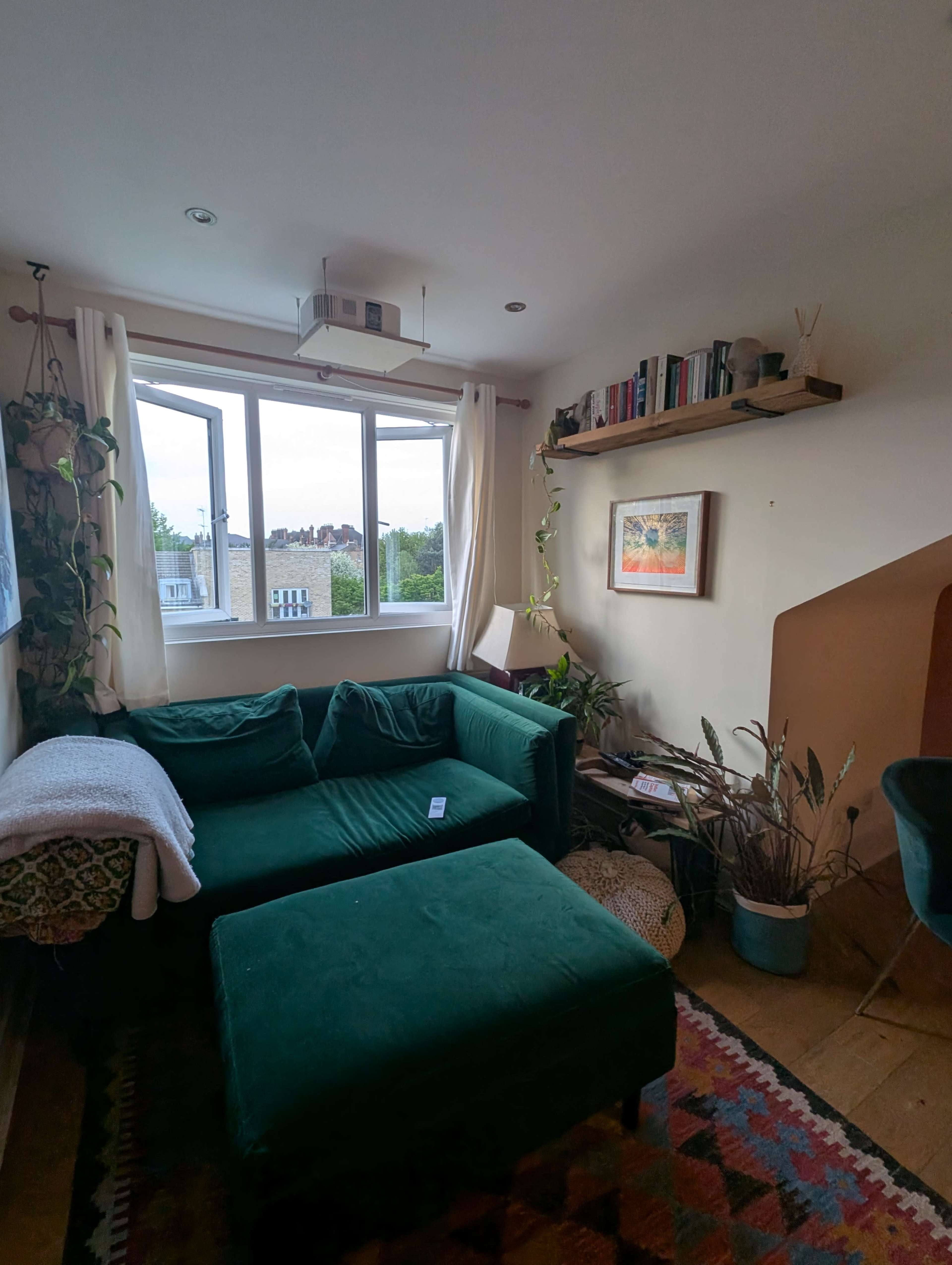 The image depicts a cozy living room with a green couch, a large window, and shelves containing books and plants.