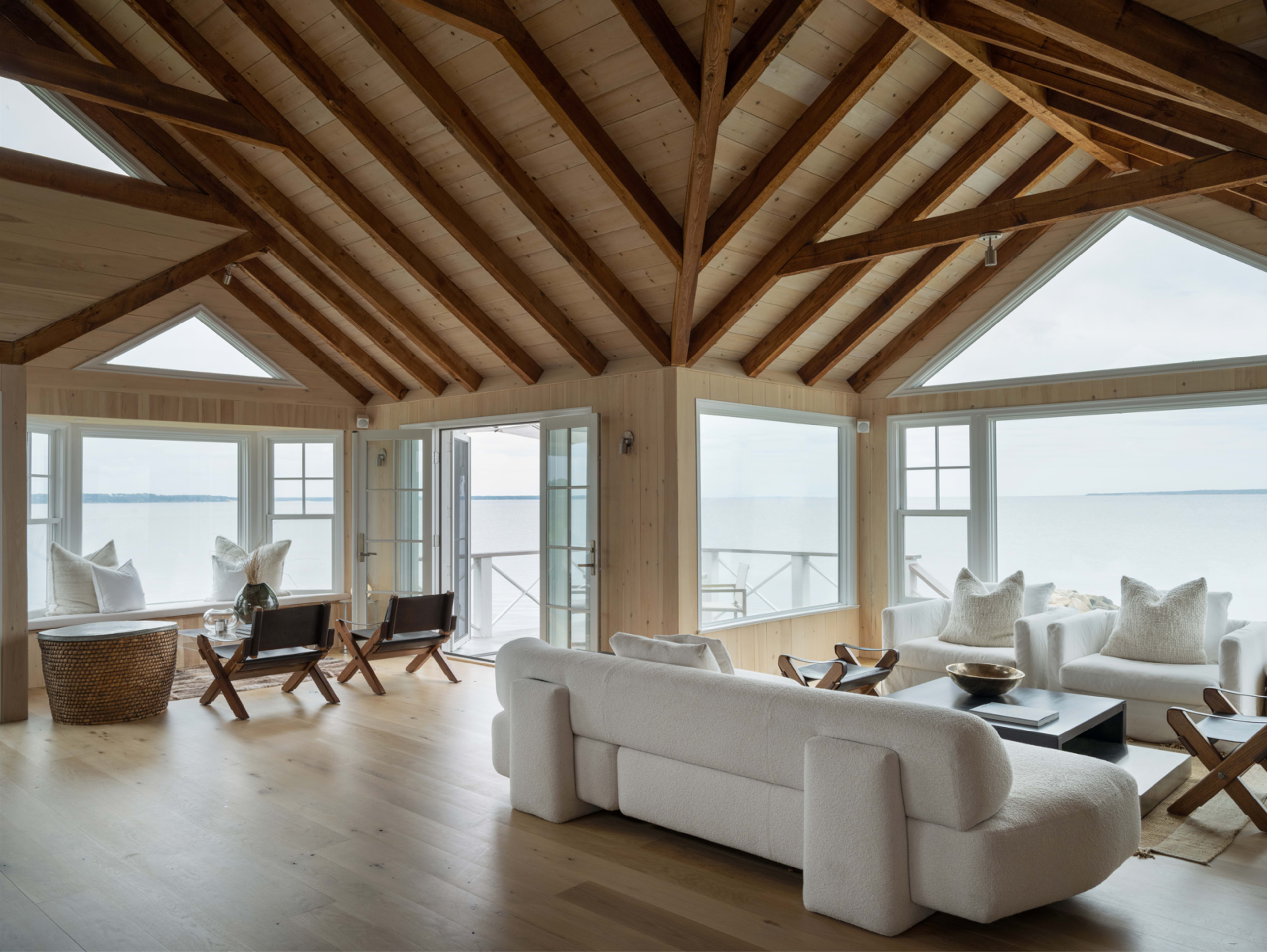 A spacious, light-filled living room with wooden beams, large windows, and a view of the water.