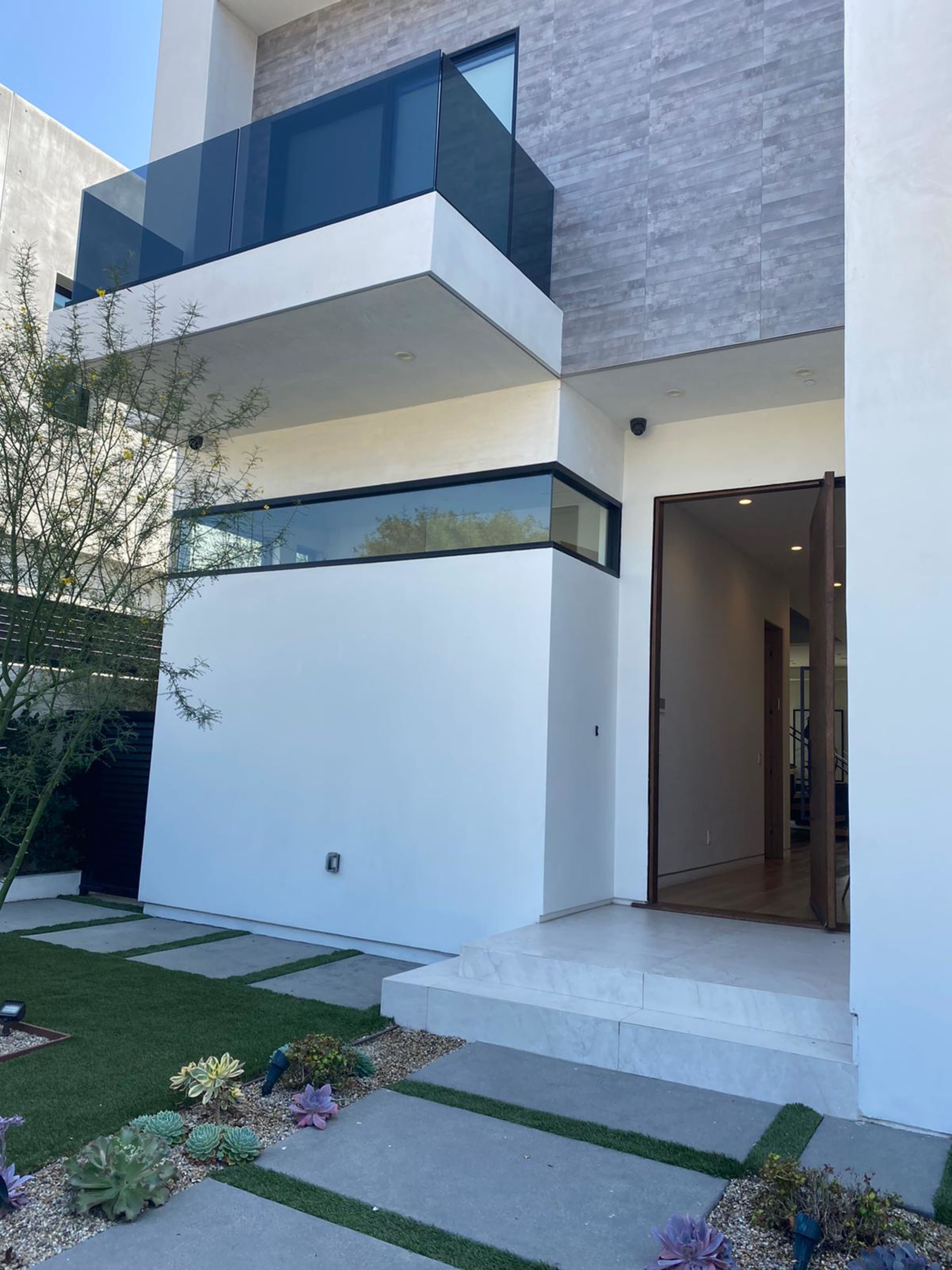 The image shows a modern two-story house with a landscaped front area featuring a path and small plants.
