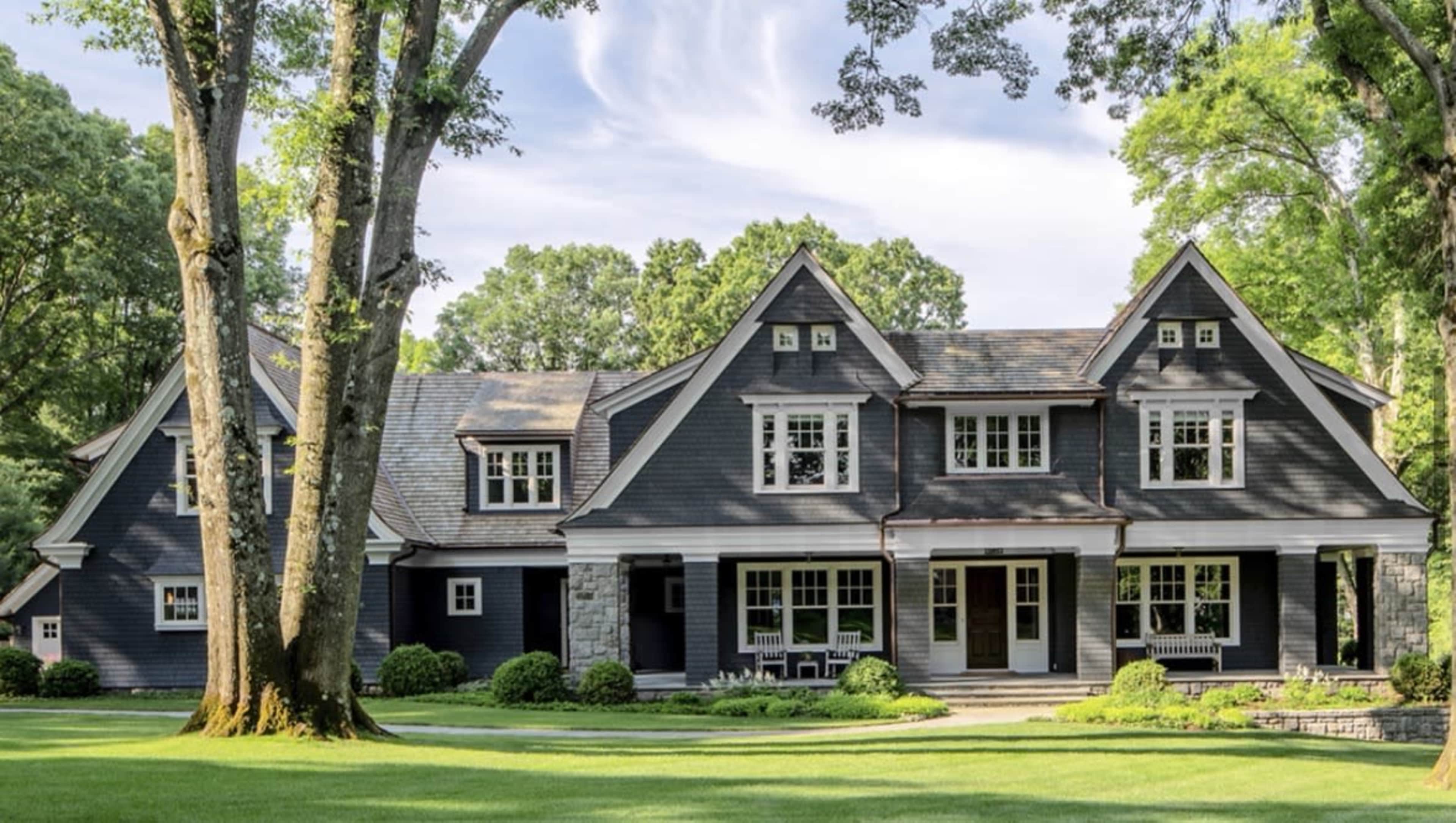 A large, dark blue house with multiple gables and a stone foundation is surrounded by green trees and a well-manicured lawn.