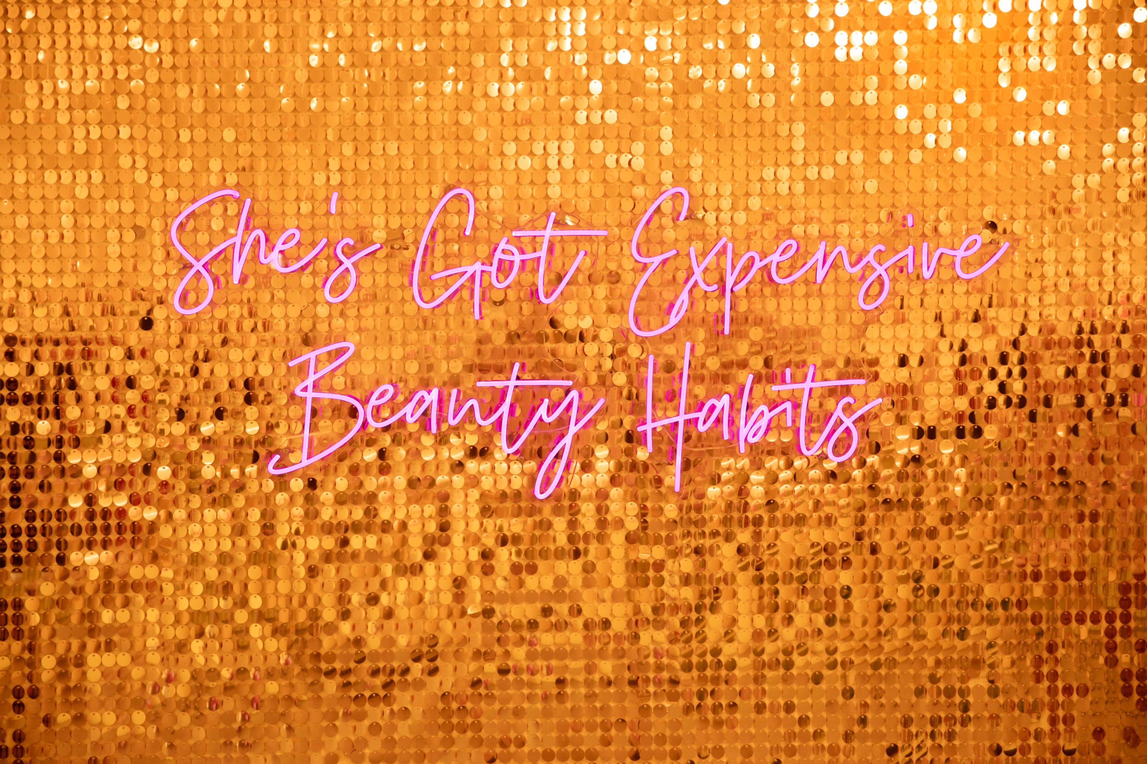 The image features a vibrant orange sequin background with the text "She's Got Expensive Beauty Habits" in pink cursive lettering.