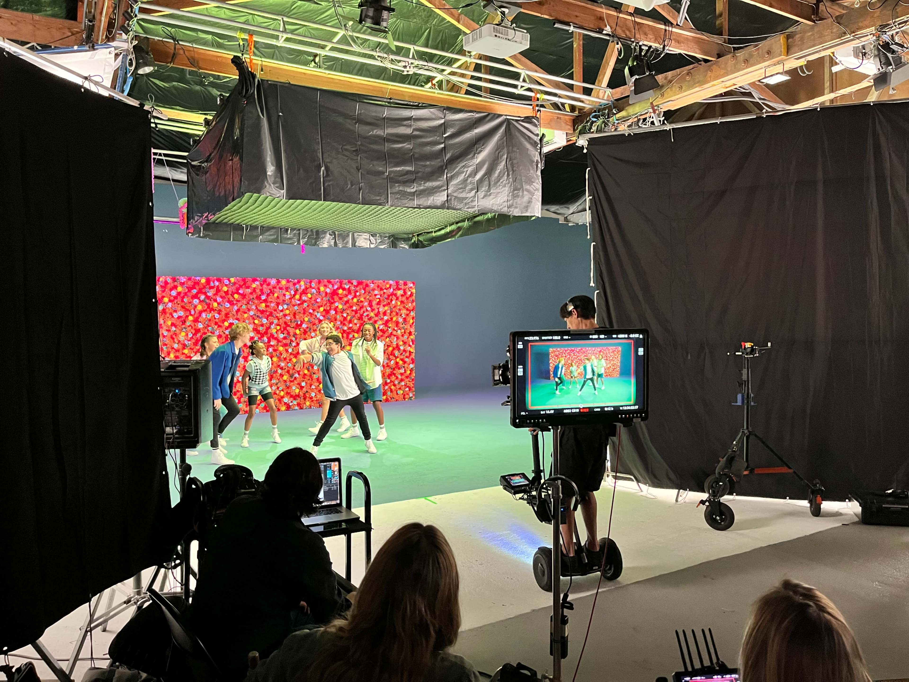 The image shows a filming studio with a colorful backdrop, where a group of dancers is performing while a crew member operates a camera on a tripod.
