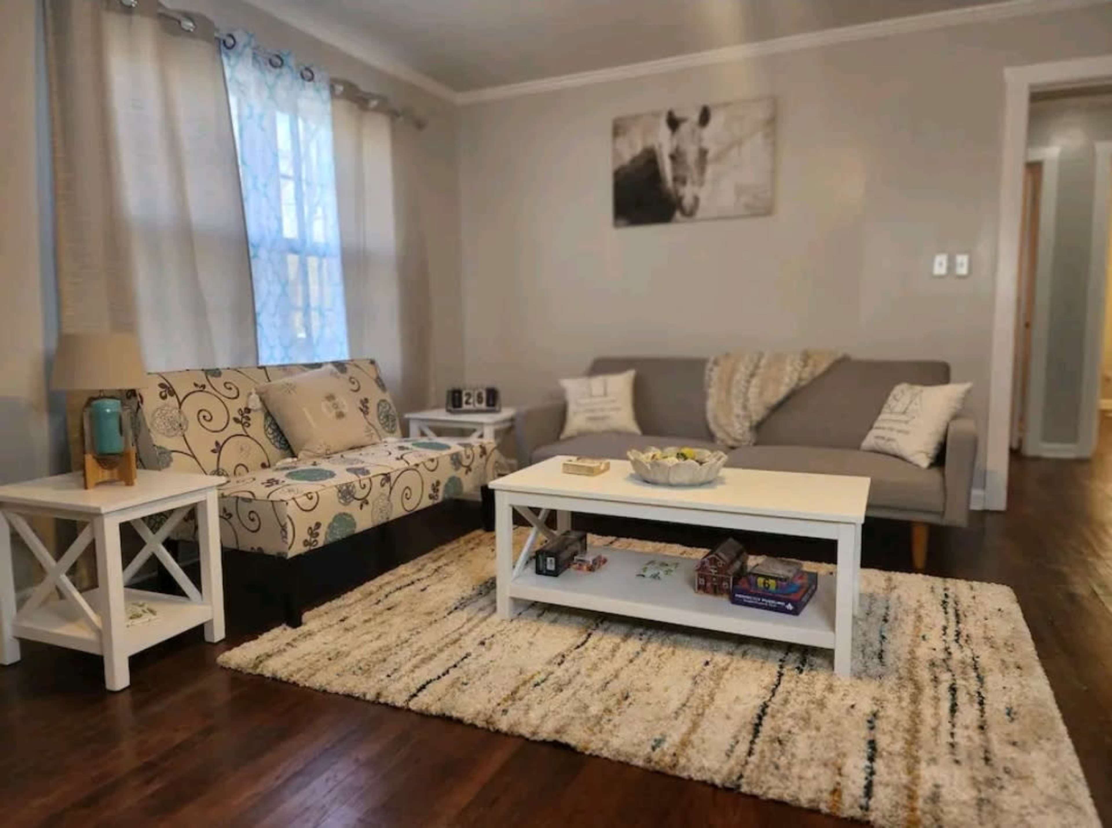 The image shows a living room with a patterned sofa, a gray couch, a white coffee table, and a large area rug on wooden flooring.