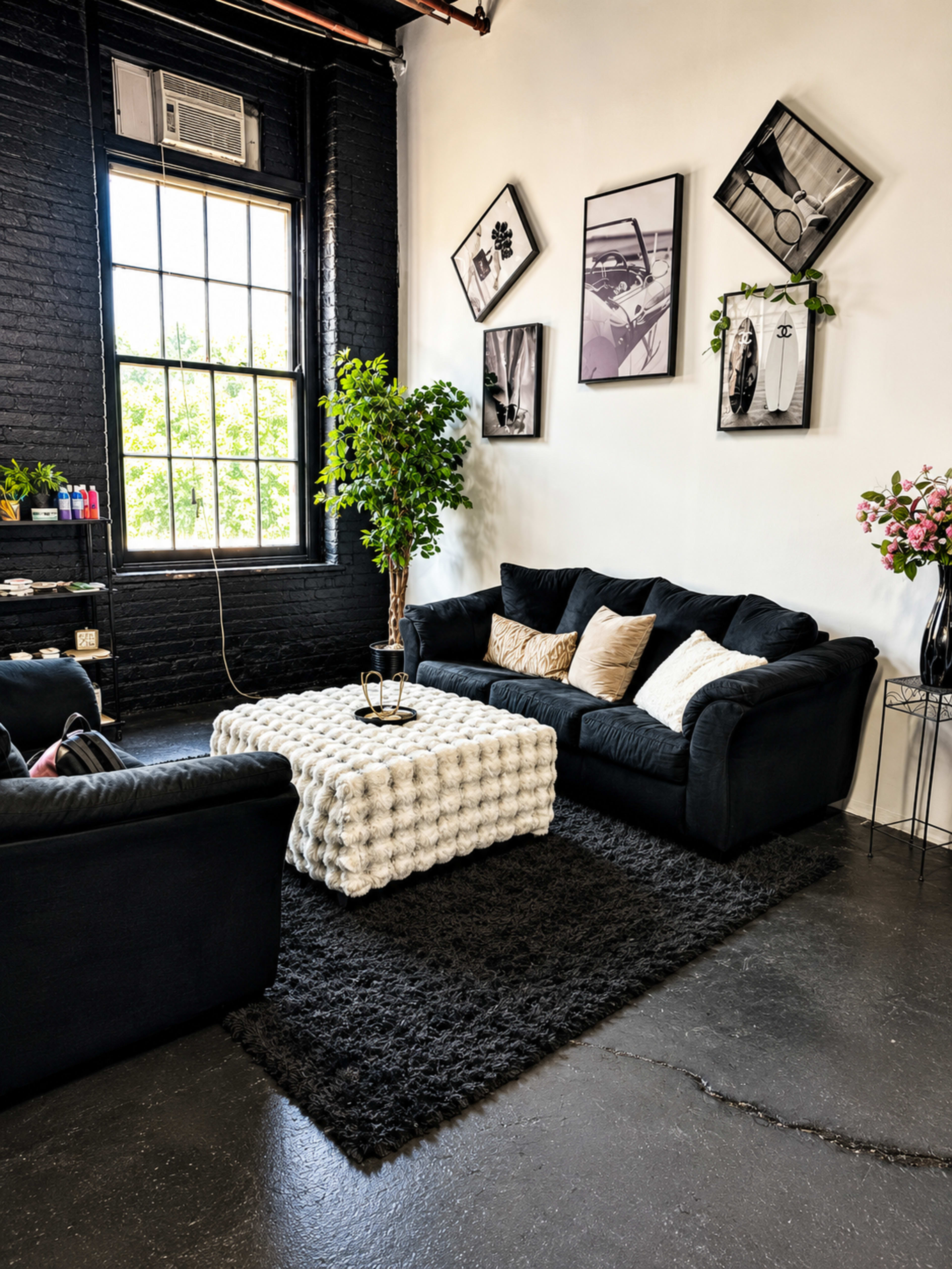 The image shows a living room with black sofas, a textured white coffee table, and framed artwork on the walls, accompanied by a green plant and floral arrangements.