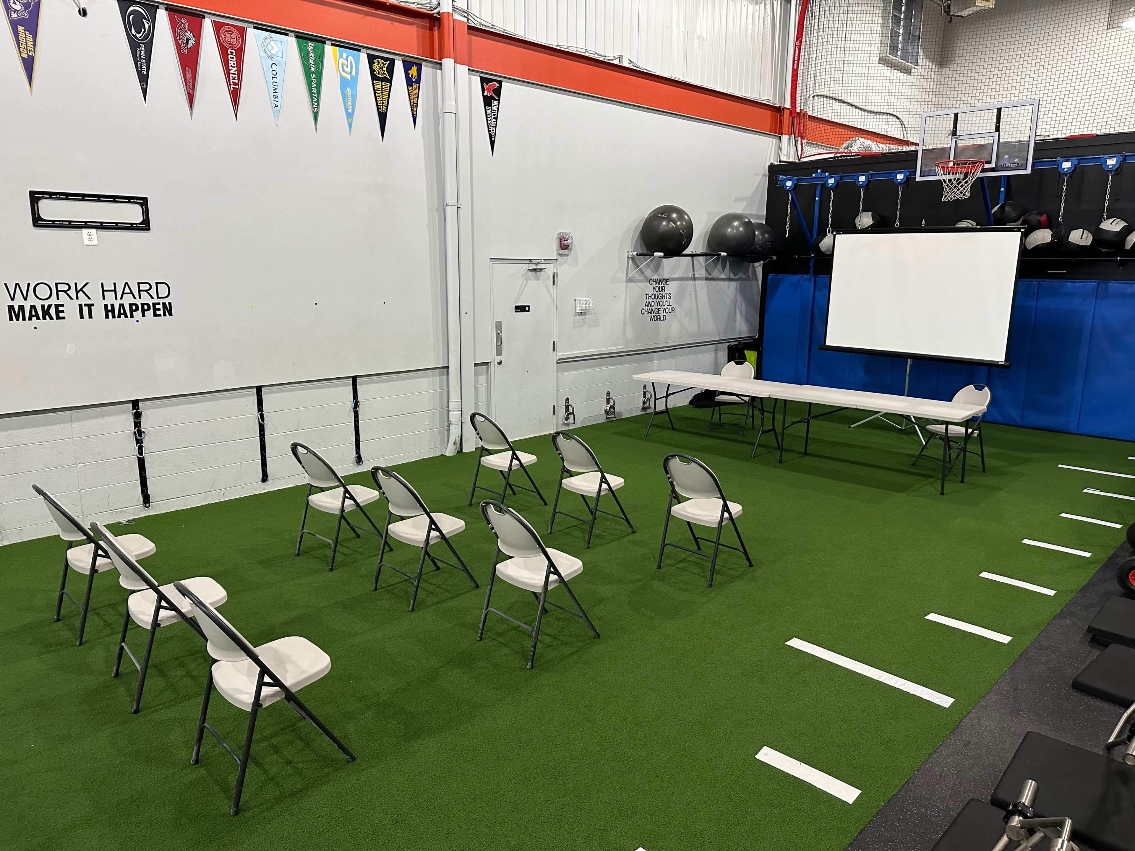 A gymnasium setup features rows of chairs facing a long table and a large screen, with exercise equipment in the background.