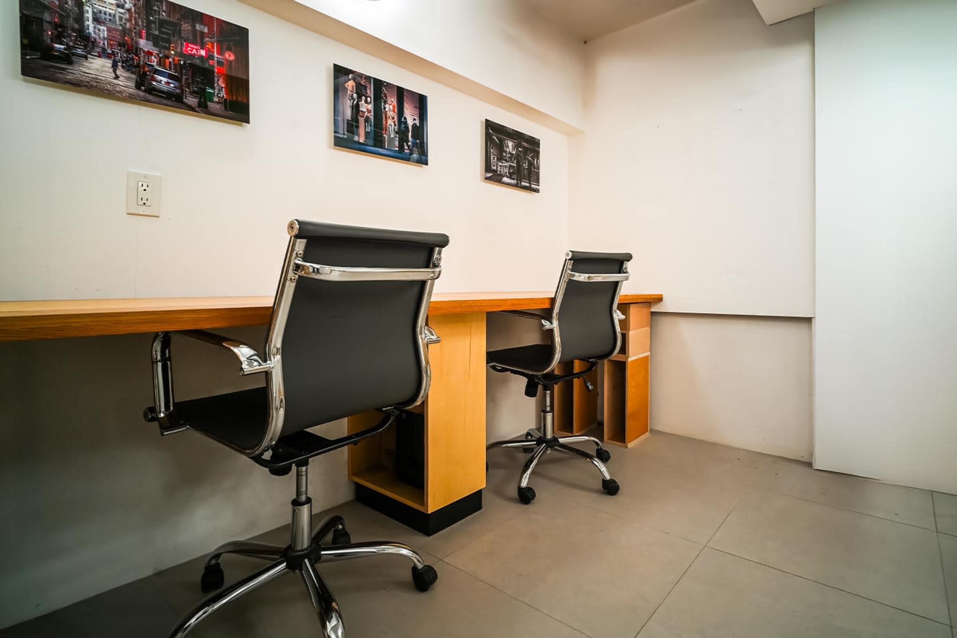 The image shows a minimalist workspace with two black office chairs and a wooden table against a white wall, featuring framed pictures above.