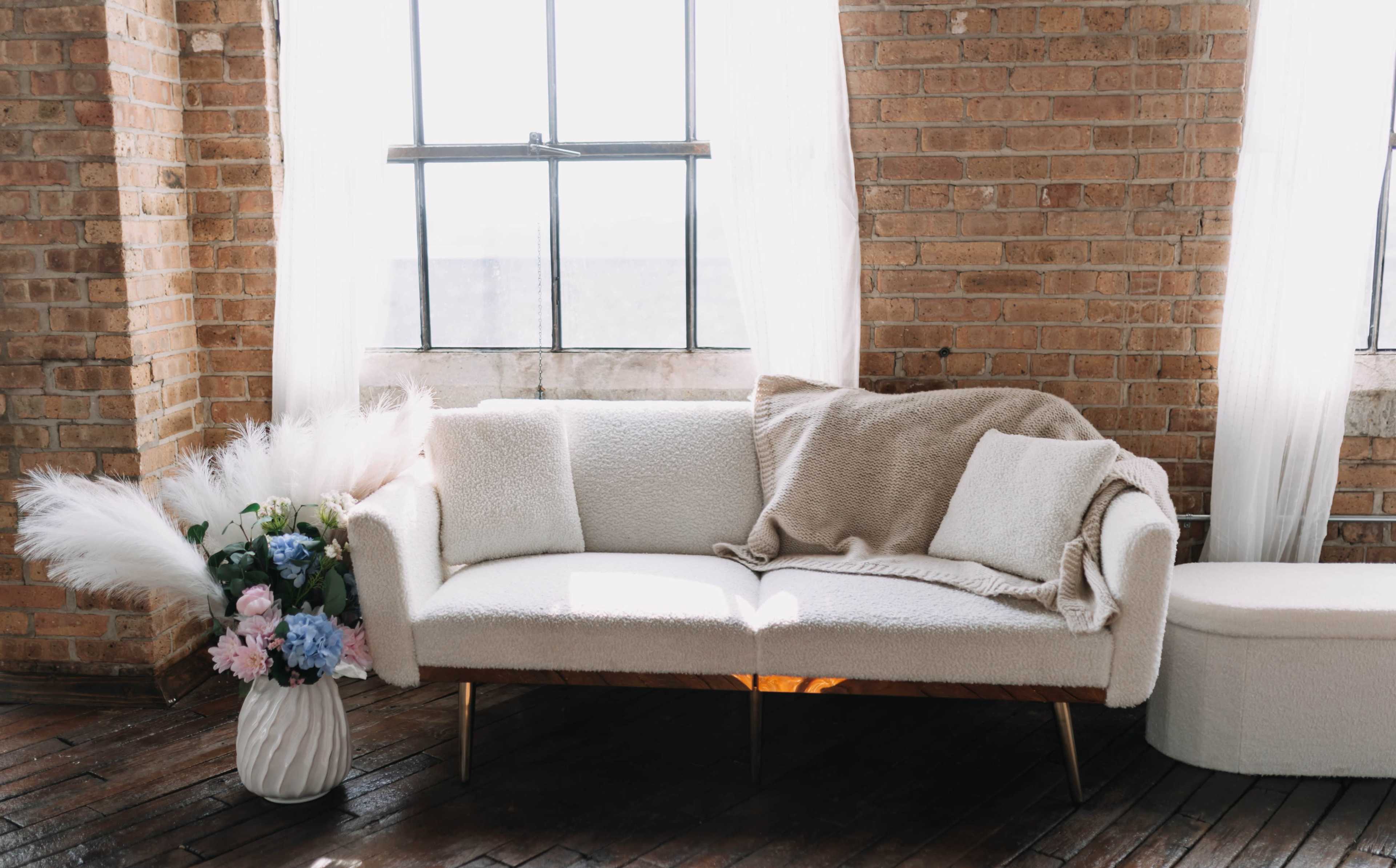 A white, textured sofa with a blanket draped over it is positioned beside a window in a room with exposed brick walls and a vase of flowers.