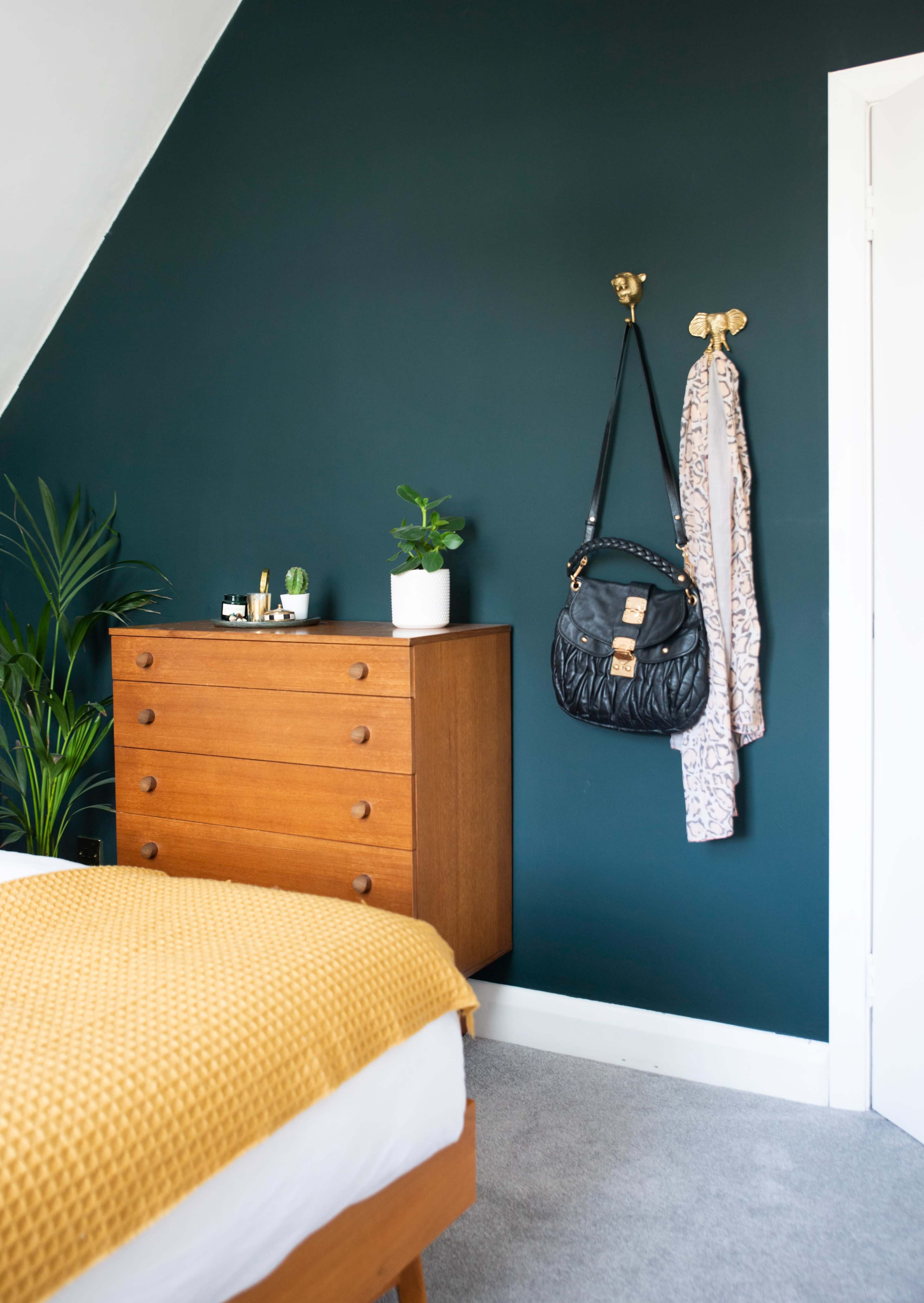 A cozy bedroom with a green accent wall, a wooden dresser, a plant in a pot, and a handbag hanging beside a garment on hooks.