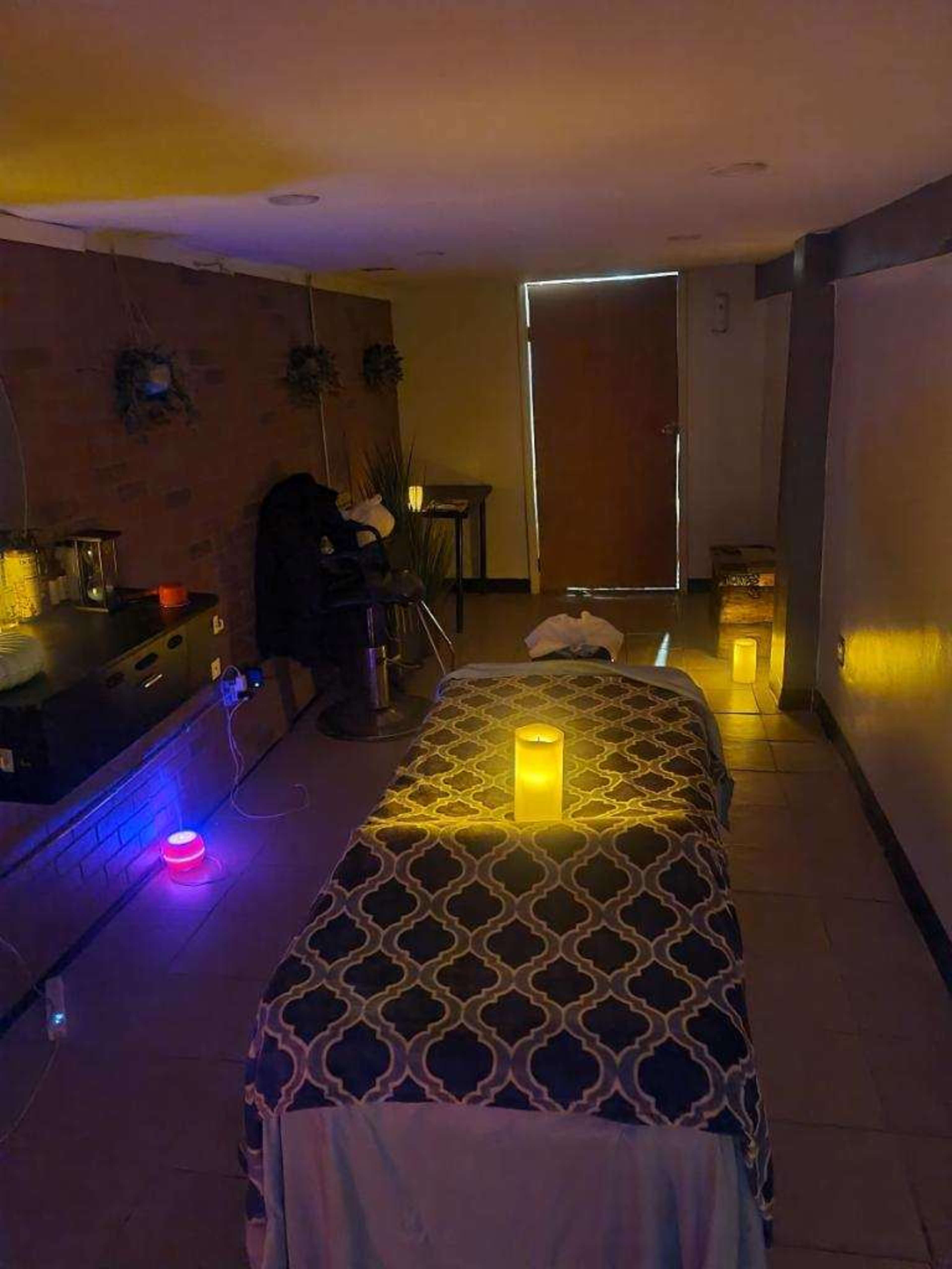 A massage room features a treatment table adorned with a patterned cover, surrounded by various glowing candles and soft lighting.