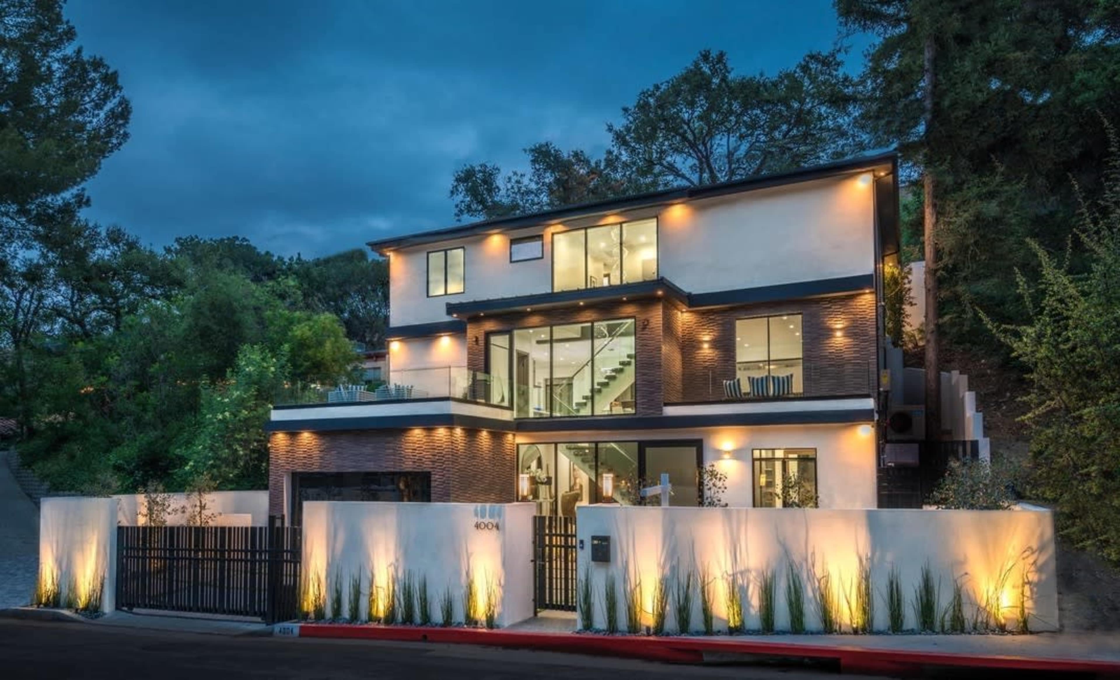 A modern, three-story house with large windows and a sleek design stands illuminated at dusk, surrounded by greenery and featuring a gated frontyard.