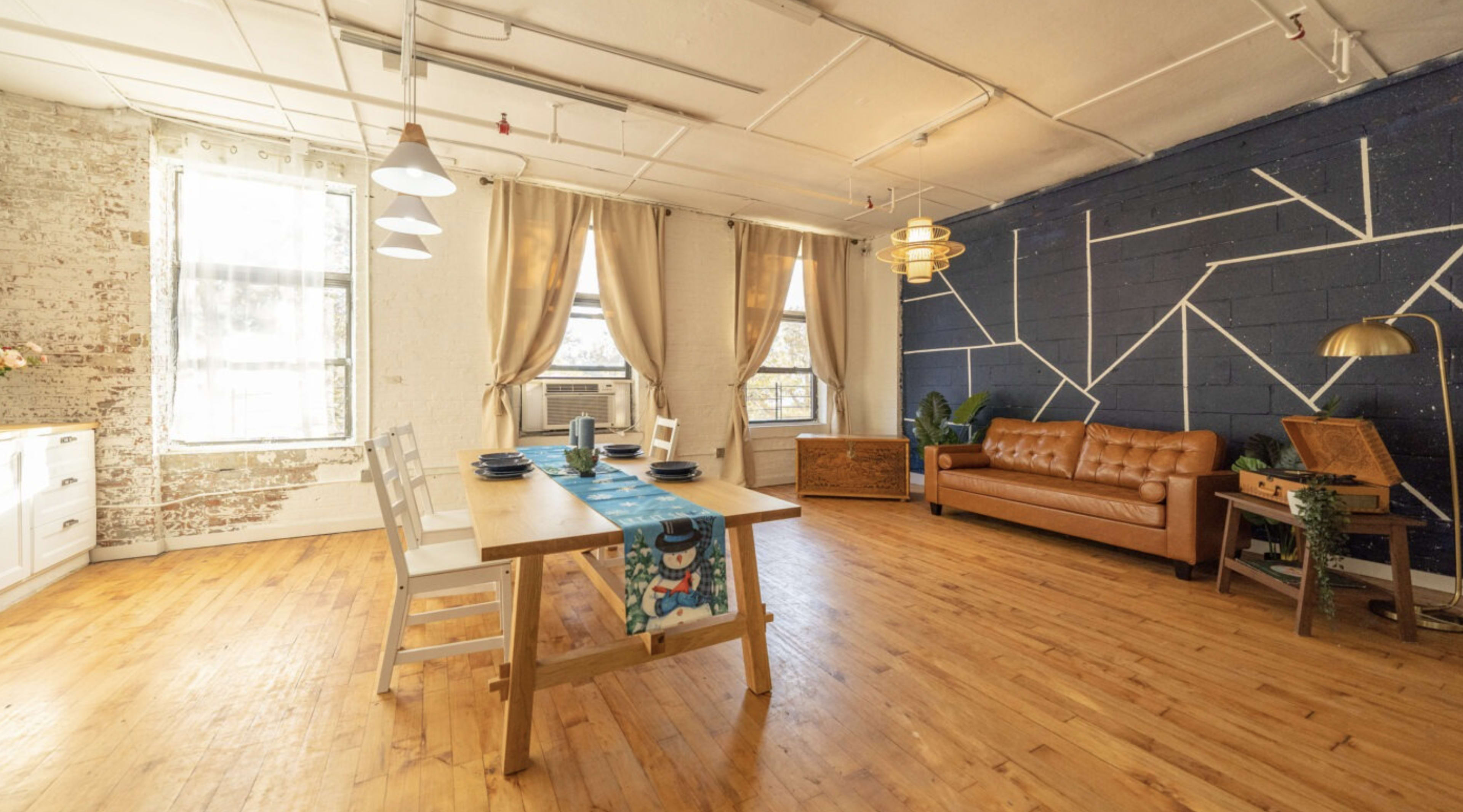 A spacious room featuring a wooden dining table with chairs, a brown leather sofa, and large windows, accented by a blue wall with geometric white lines.