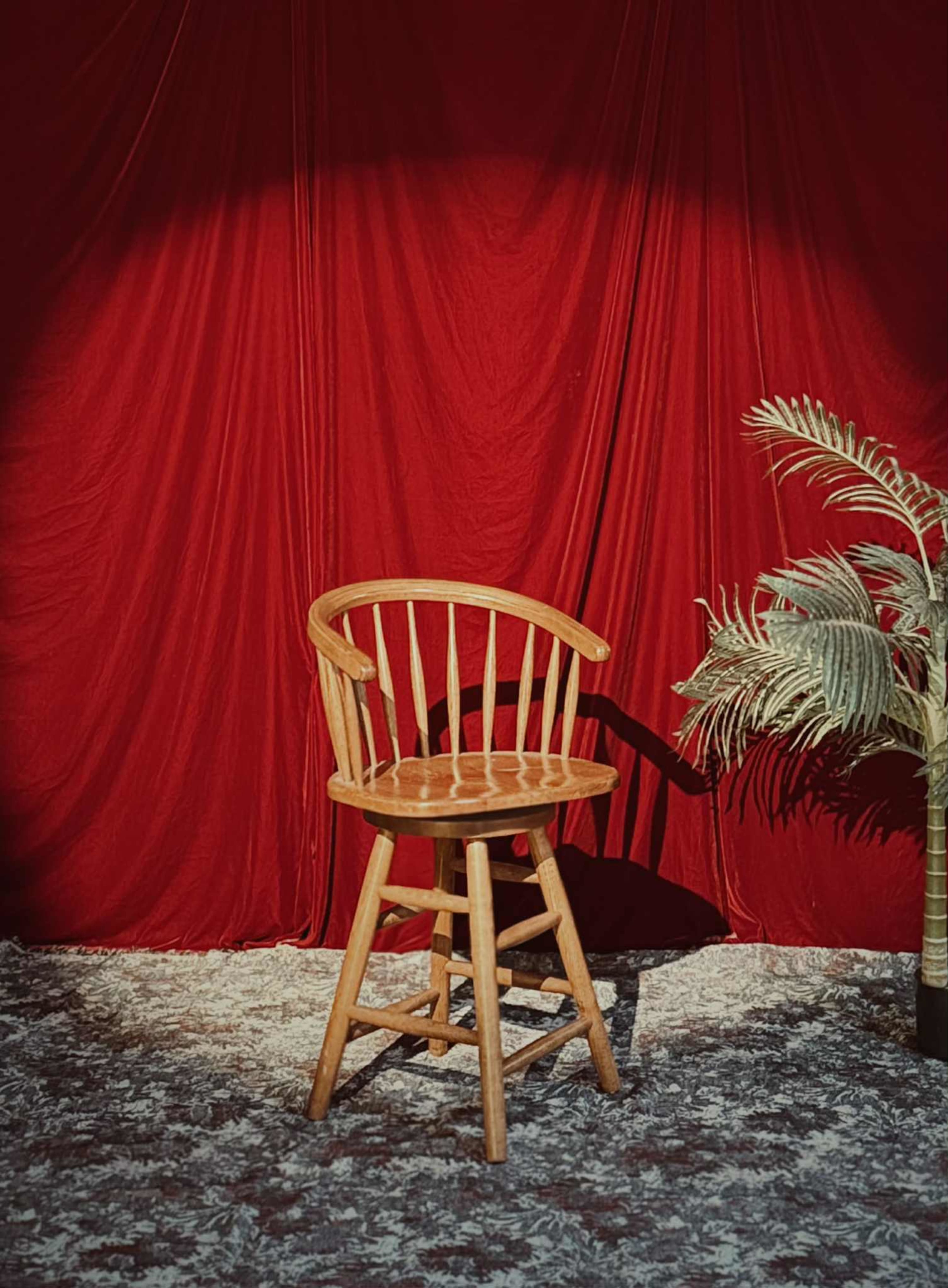 A wooden chair with a curved backrest is positioned in front of a red curtain, alongside a potted plant.