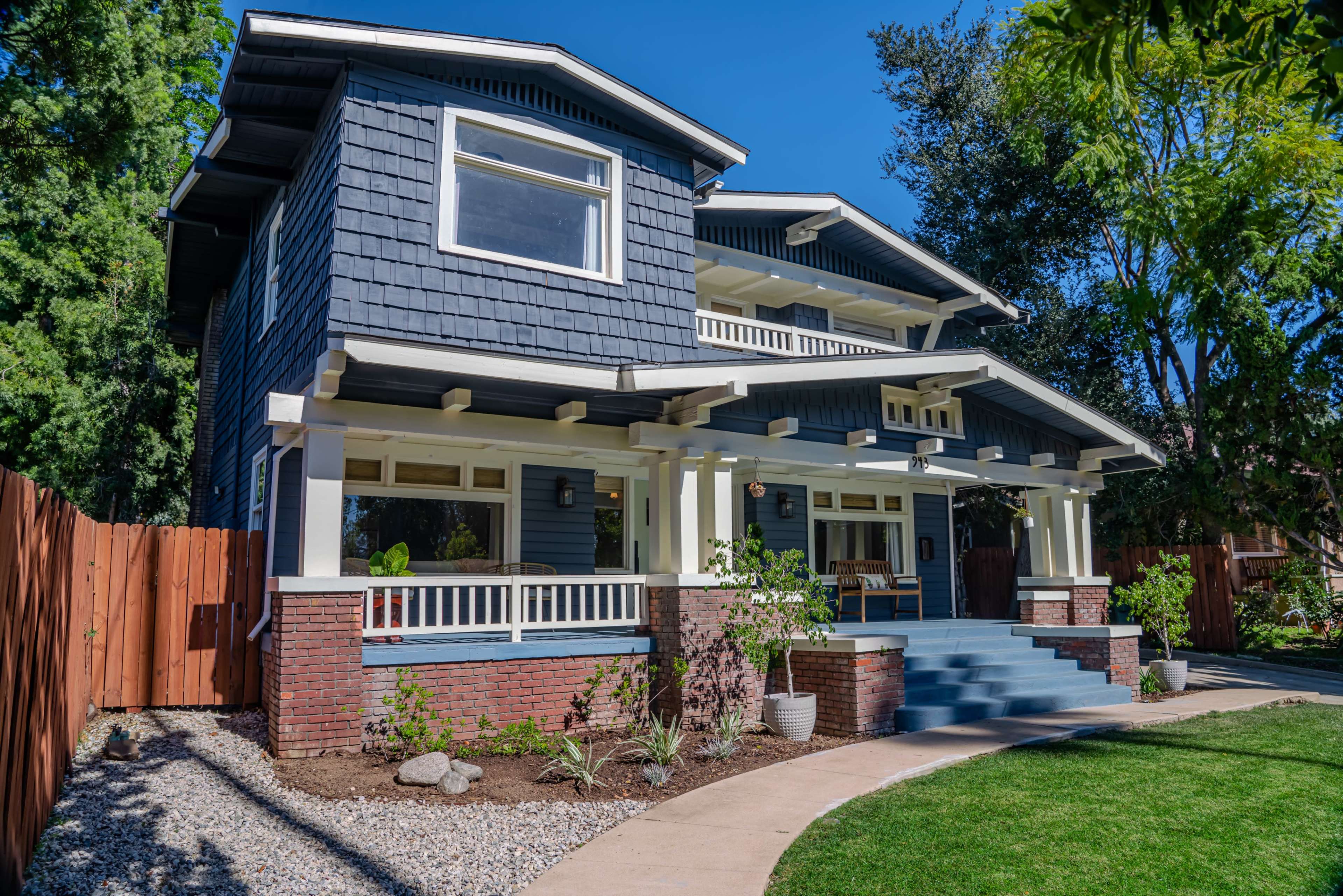 A two-story blue house with a wraparound porch, surrounded by a landscaped yard and wooden fence.