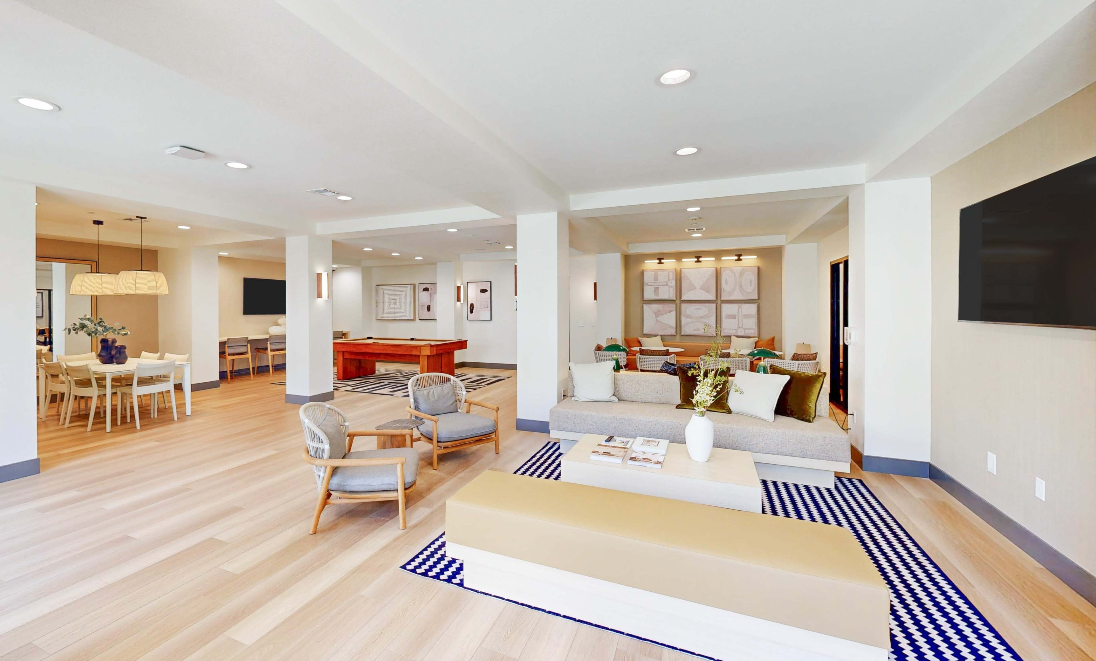 The image shows a modern open-concept lounge area featuring comfortable seating, a dining space, and a polished wooden table in a well-lit environment.