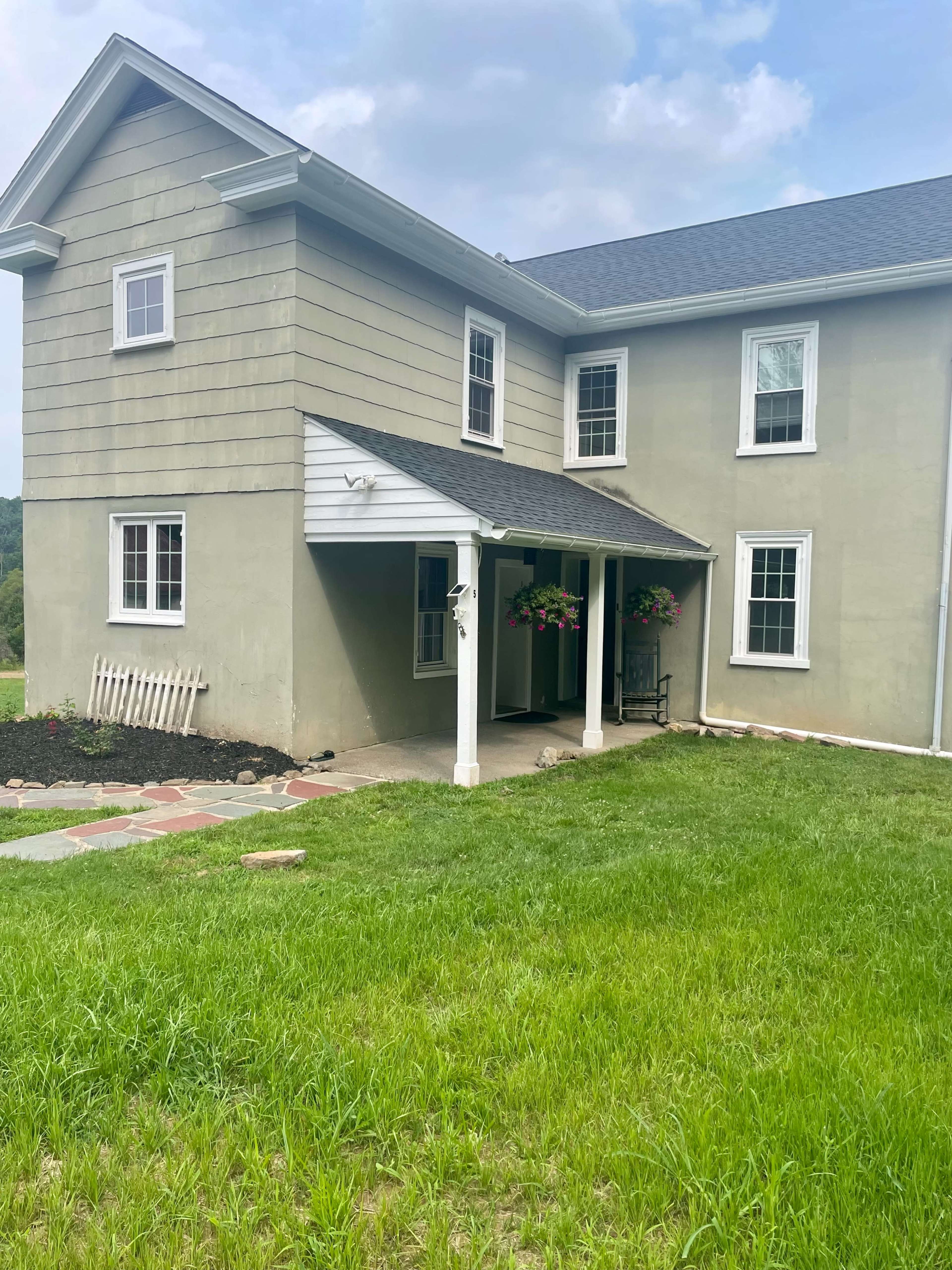 A two-story house with a green exterior, multiple windows, a front porch, and hanging flower baskets is set against a grassy yard.