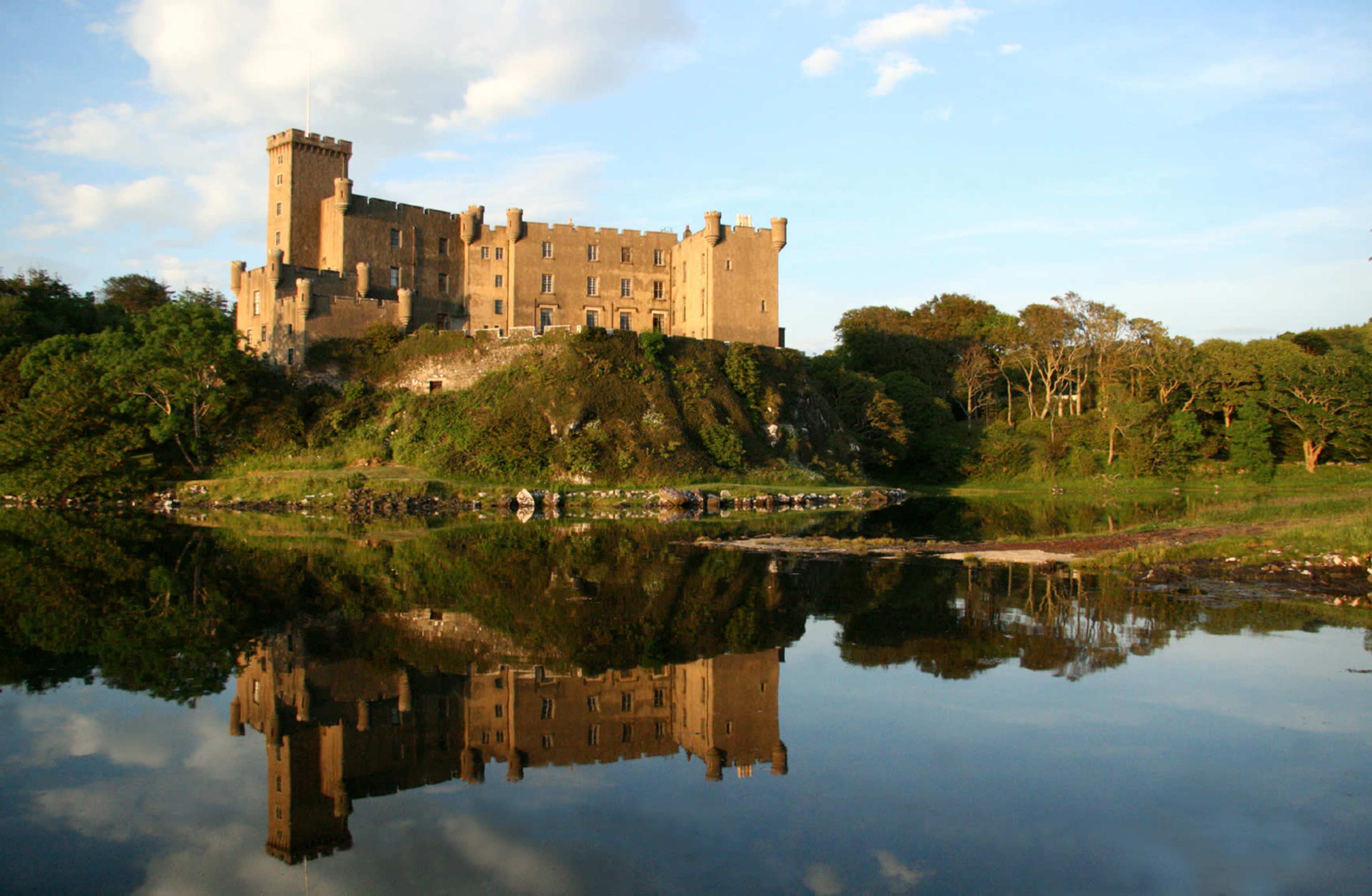 A stone castle stands on a grassy hilltop, reflecting in the calm waters of a nearby lake, under a blue sky with scattered clouds.