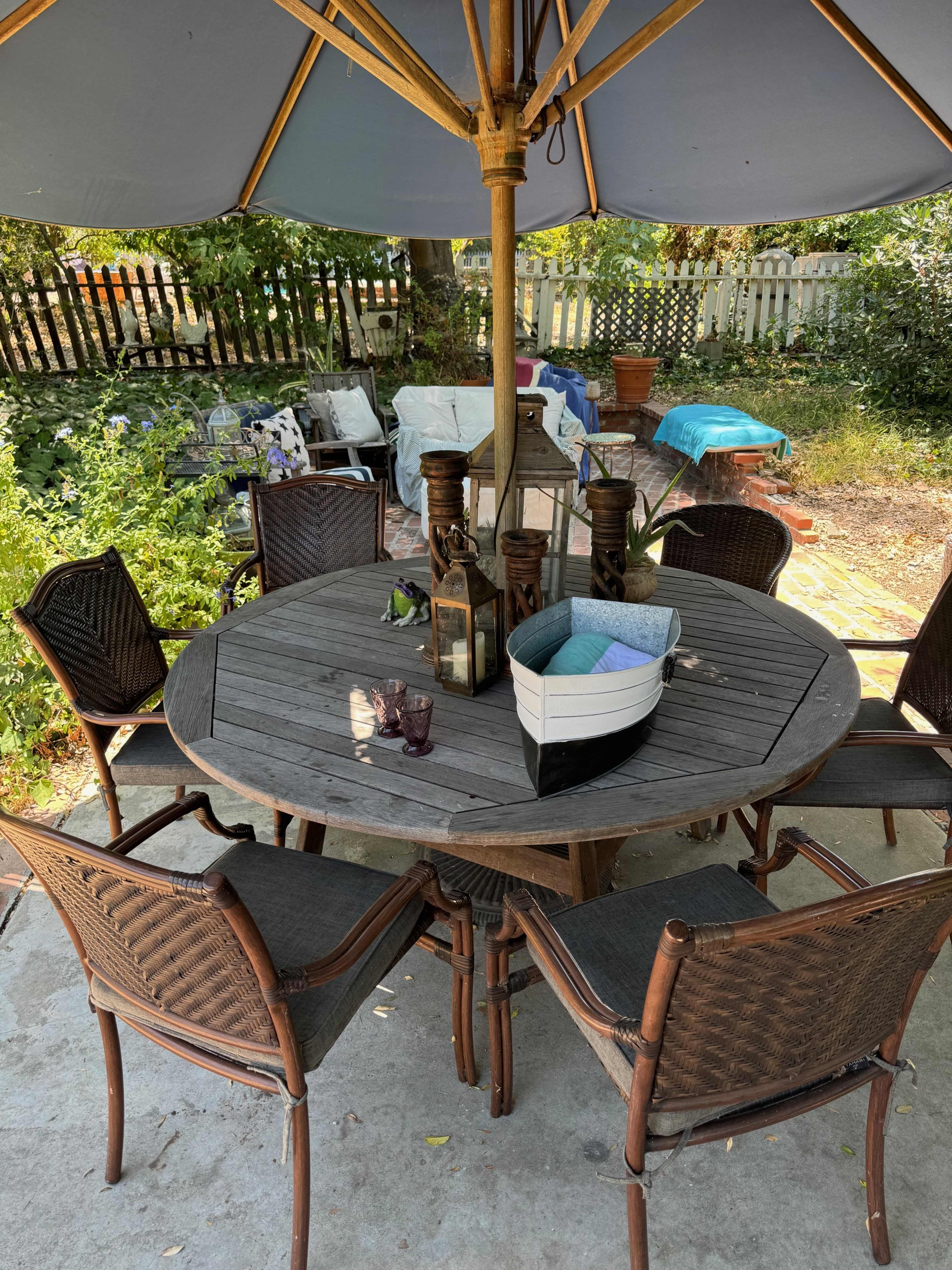 A round wooden dining table with six chairs is set beneath a large umbrella in a sunny outdoor space surrounded by greenery and garden furniture.