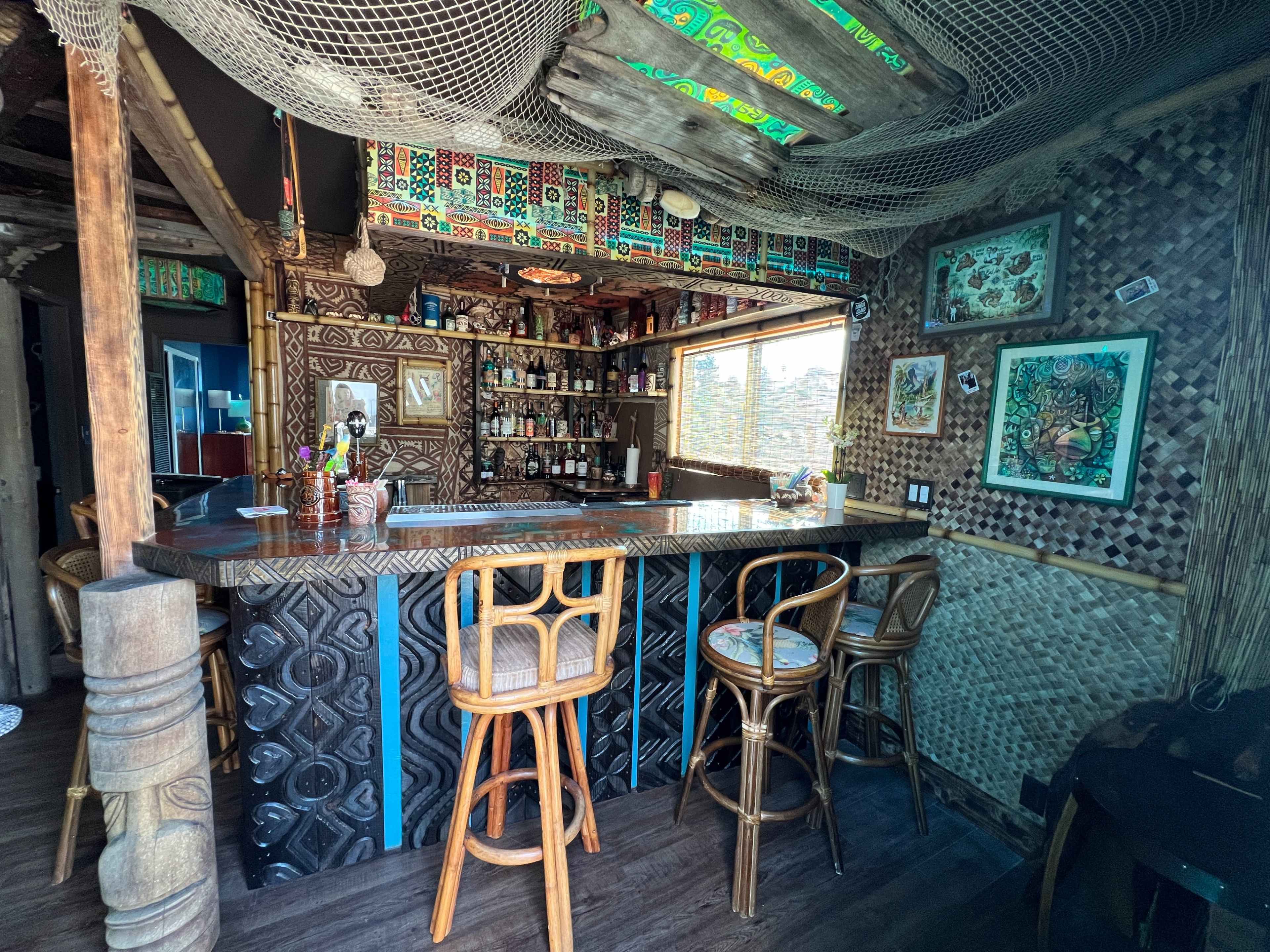 A rustic bar area with a wooden counter, woven decorations, and shelves stocked with bottles, set in a well-lit room featuring large windows.