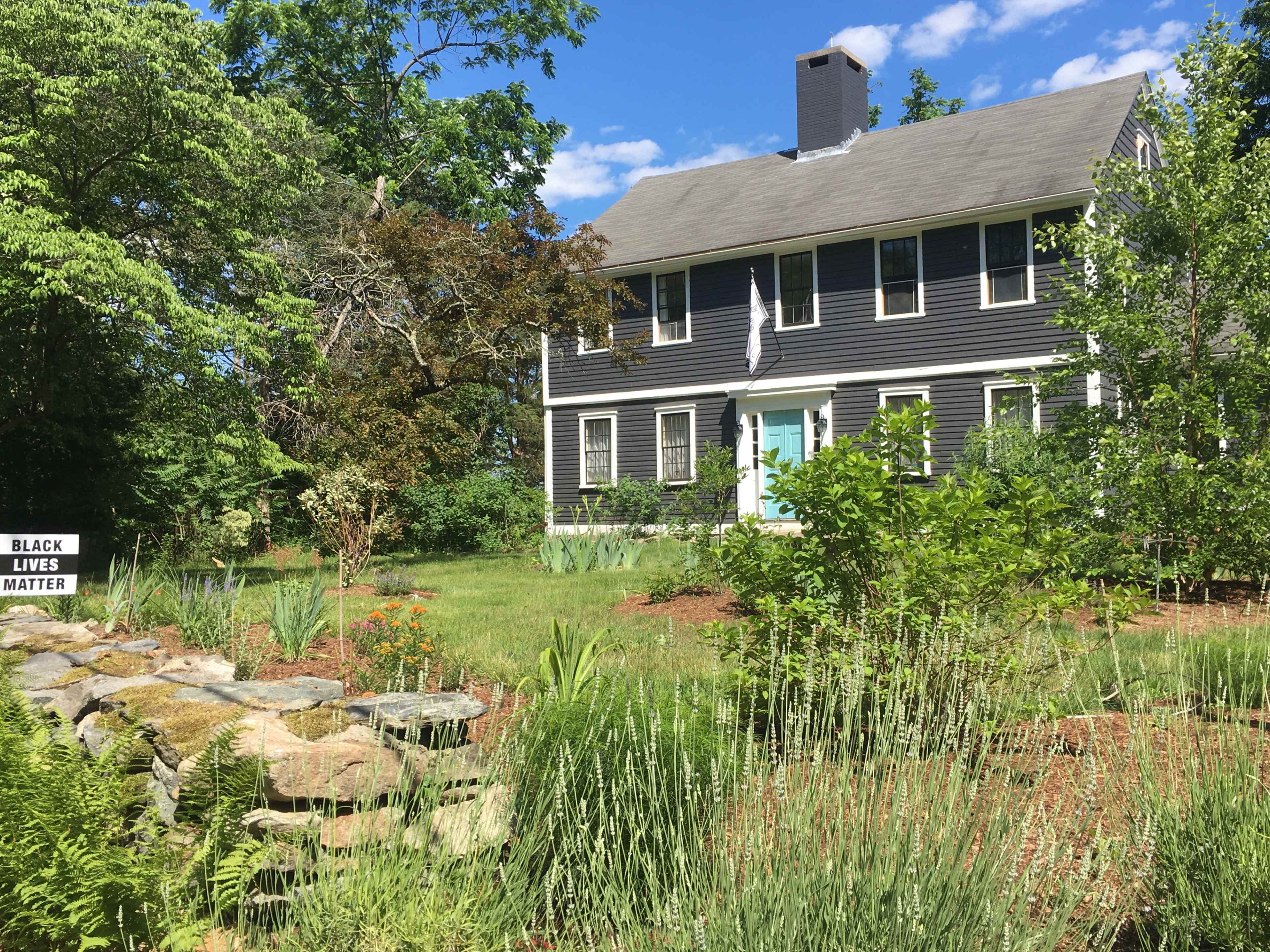 A two-story gray house with a blue front door is surrounded by a garden featuring various green plants and a sign reading "BLACK LIVES MATTER" in the yard.