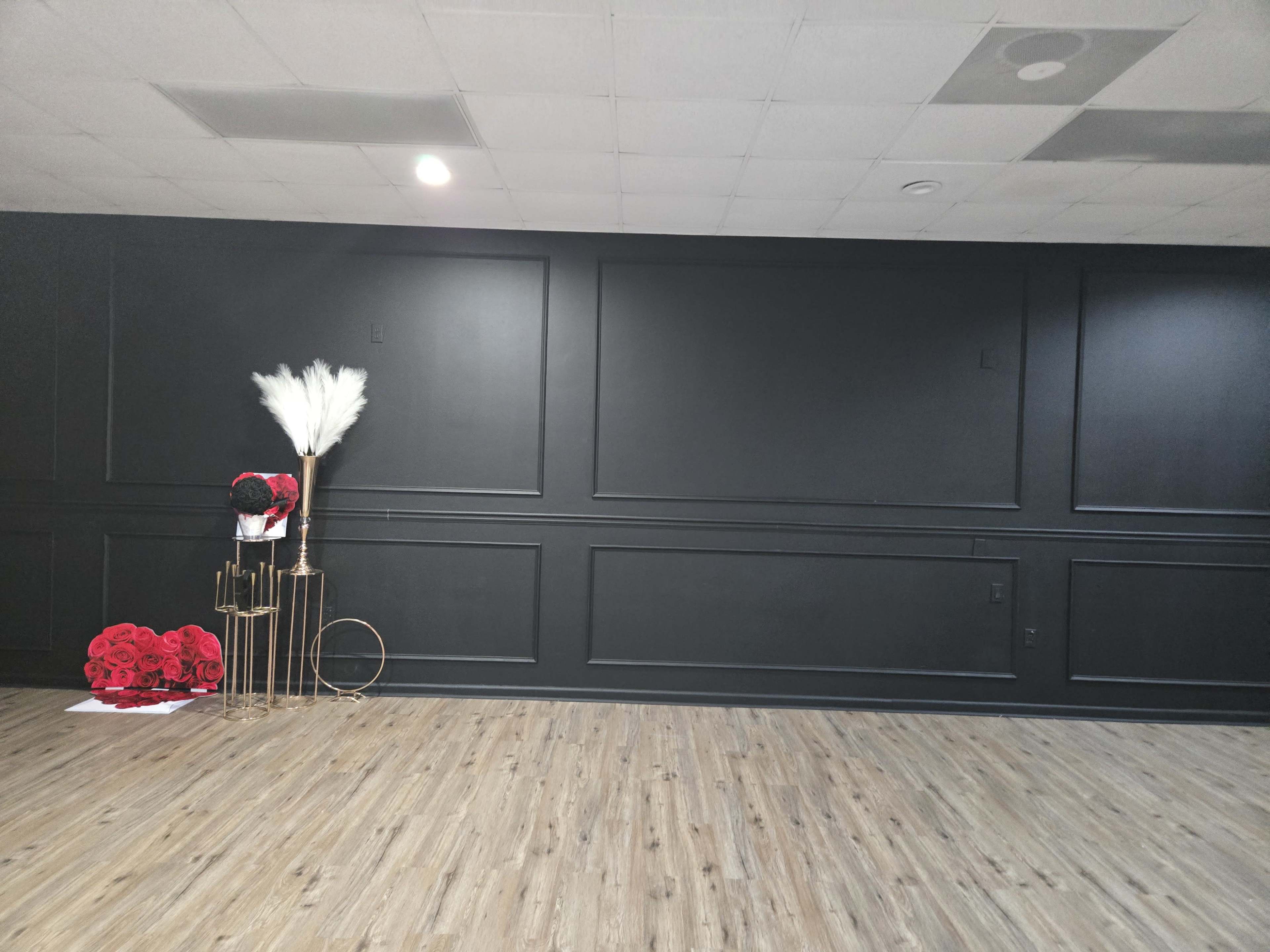The image shows a minimalist black room with a wooden floor and a decorative arrangement of red flowers and a pampas grass bouquet in a gold stand against the wall.