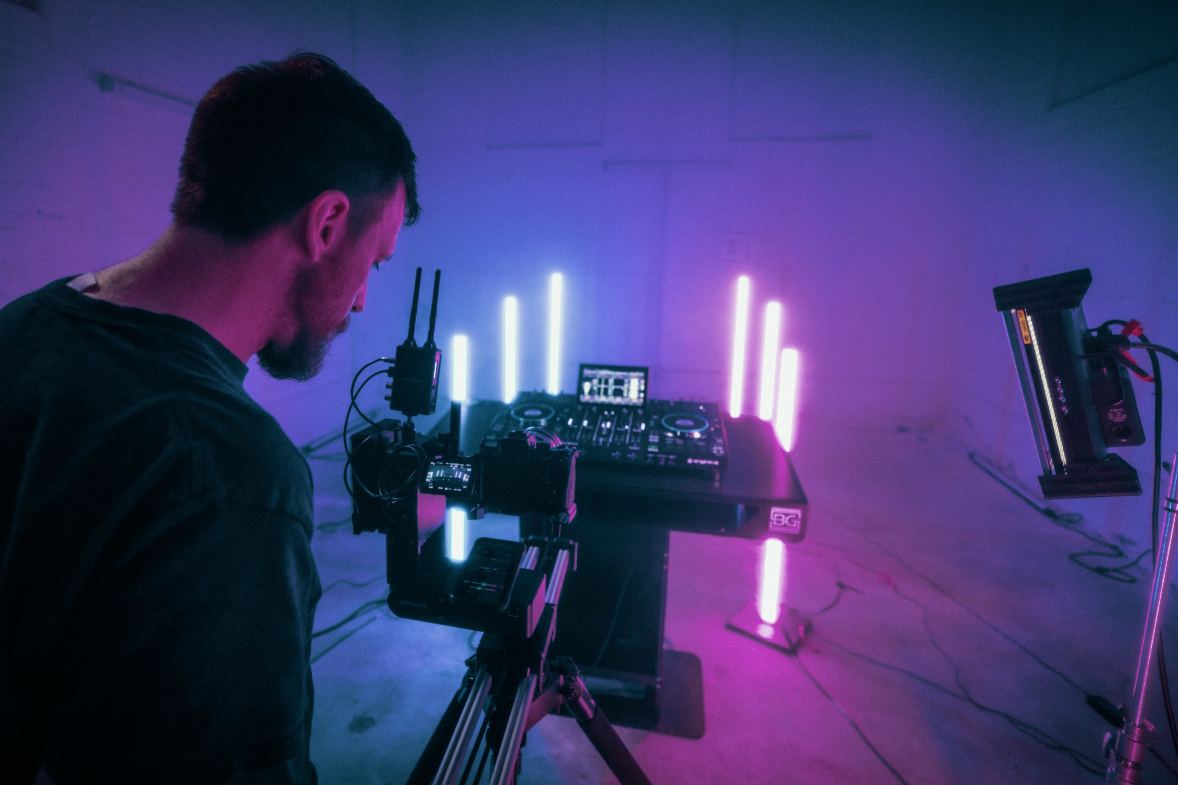 A man sets up a camera in front of a DJ station illuminated by colorful lights in a dimly lit room.